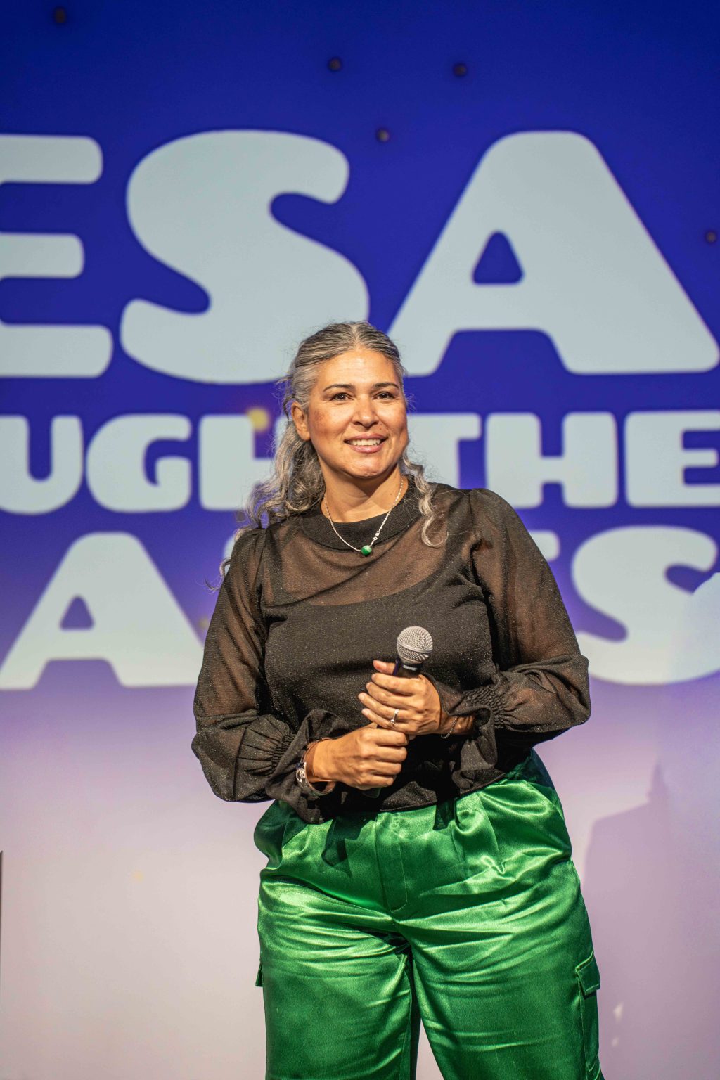 A woman with gray hair wearing a sheer black blouse and shiny green pants holds a microphone and smiles on stage in front of the MESA Through the Decades 2025 Photo Gallery blue and white backdrop with large partial letters.