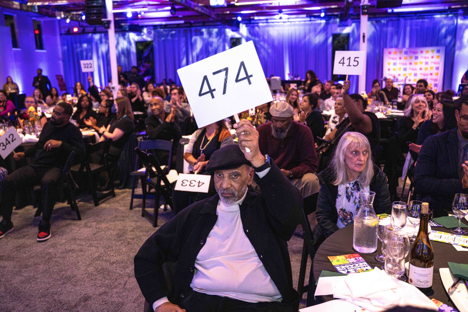 At the MESA Through the Decades 2025 Photo Gallery, a man sitting at a table raises a sign with the number 474, surrounded by other attendees holding numbered signs in a purple-lit room.