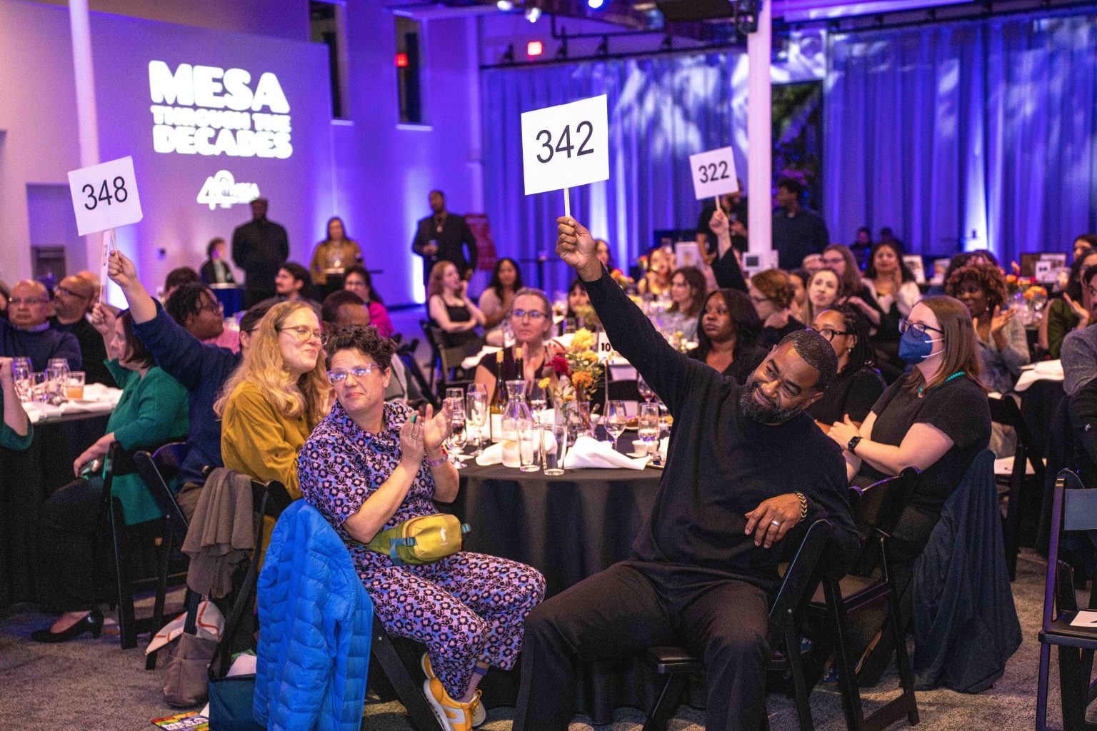 A lively auction scene at the MESA Through the Decades 2025 Photo Gallery gala, with attendees seated at round tables. Several people are raising numbered paddles, including a man holding up 342. The room is warmly lit with purple and blue accents.