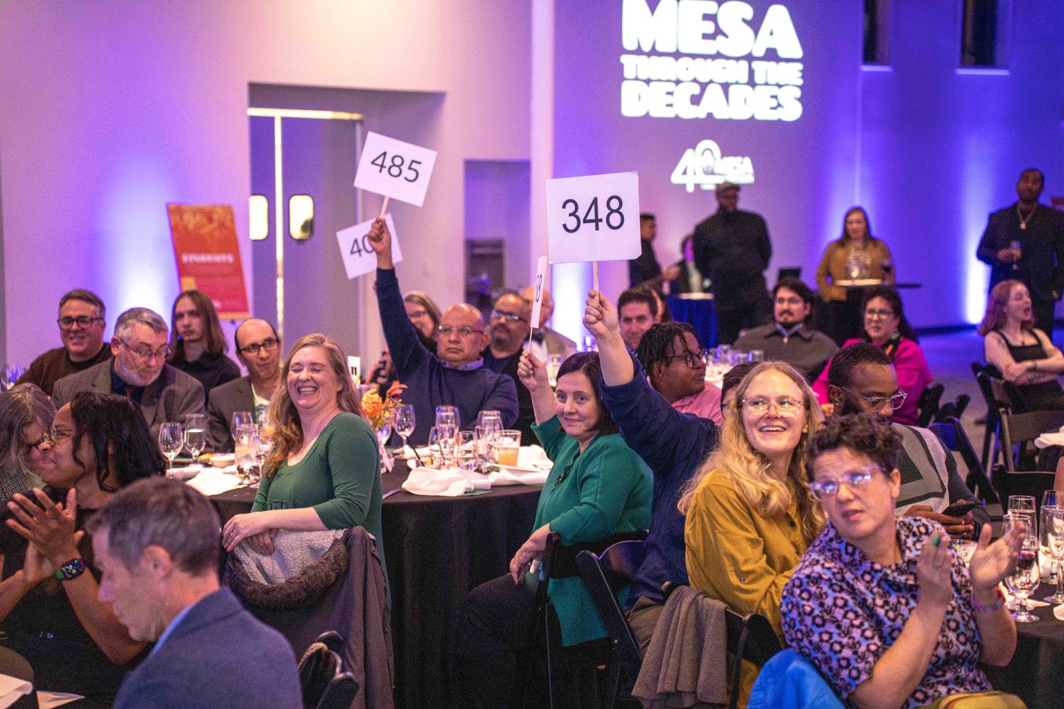 A group of people seated at round tables during an indoor event, some holding up numbered bidding paddles. The room is lit with purple light, and a wall display reads MESA THROUGH THE DECADES. Perfect for the MESA Through the Decades 2025 Photo Gallery.