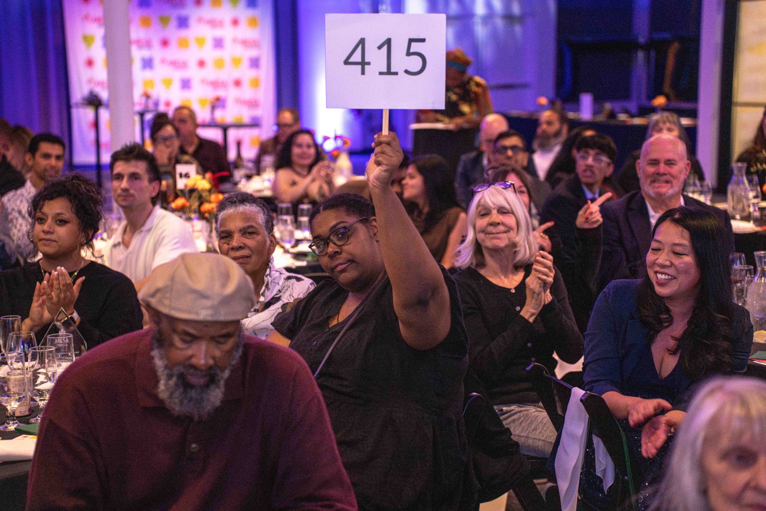 A woman holds up a sign with 415 at the lively MESA Through the Decades 2025 Photo Gallery event, while others around her sit at tables, clapping and watching attentively. The atmosphere appears engaged and celebratory.