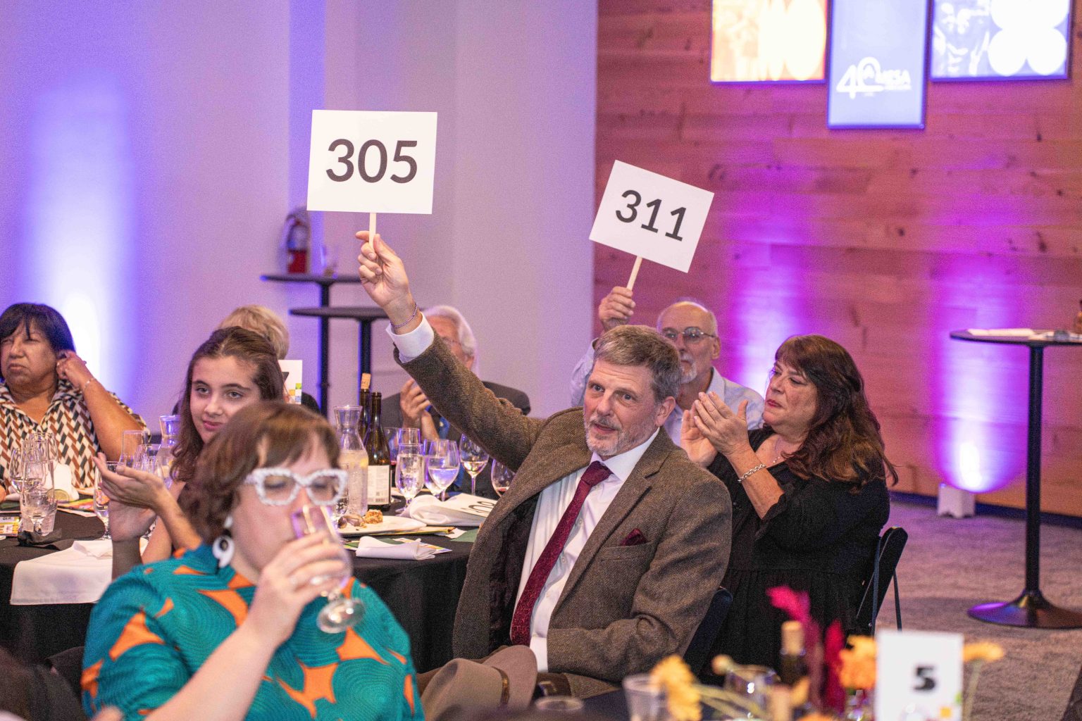 At the MESA Through the Decades 2025 Photo Gallery, people seated at tables enjoy a lively atmosphere. A man in a suit holds up a 305 sign while another person raises 311, as others clap or watch in the warmly lit room.