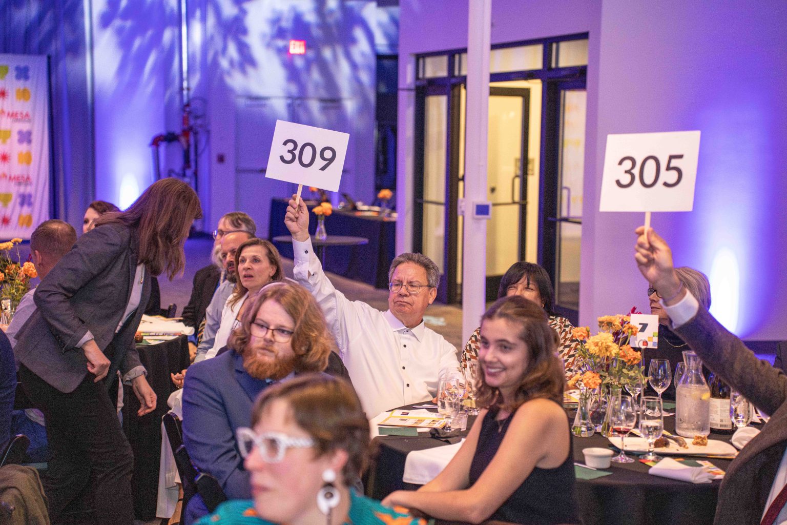 People at a formal event sit at round tables, some holding up numbered bidding paddles (309 and 305) during an auction. The lively scene, captured in the MESA Through the Decades 2025 Photo Gallery, features blue lighting and floral centerpieces.