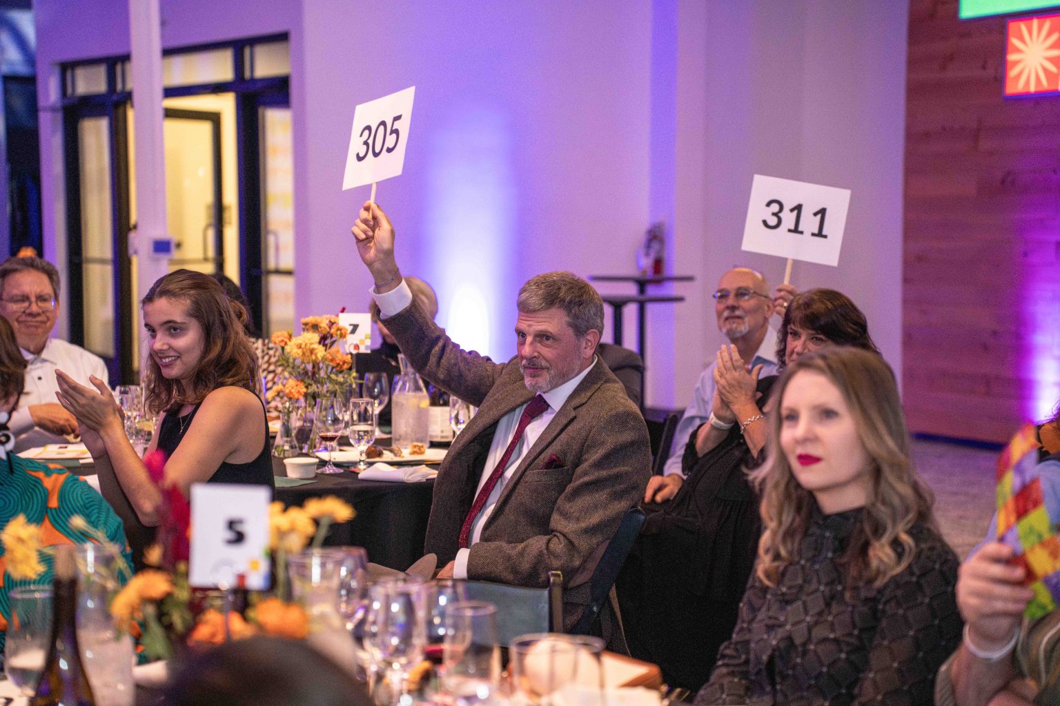 People seated at tables during the MESA Through the Decades 2025 Photo Gallery event. A man in a suit holds up a paddle with 305, while another holds 311. Others are clapping or watching amid flowers, drinks, and number cards on the tables.