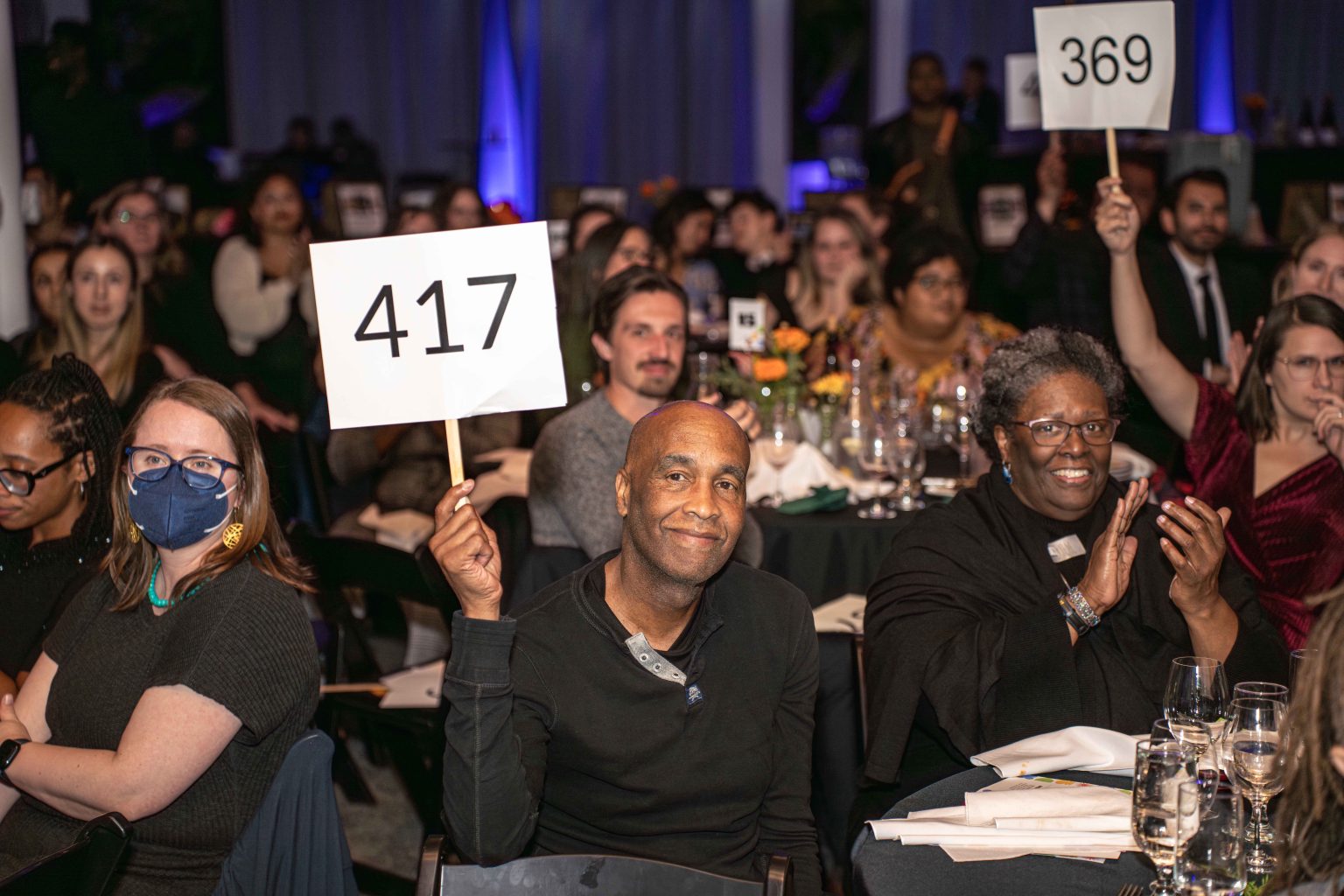 At the MESA Through the Decades 2025 Photo Gallery, a man at an event holds up a sign with the number 417, while people around him clap and another holds a sign with 369. The audience sits at tables with drinks and flowers, some smiling and watching.