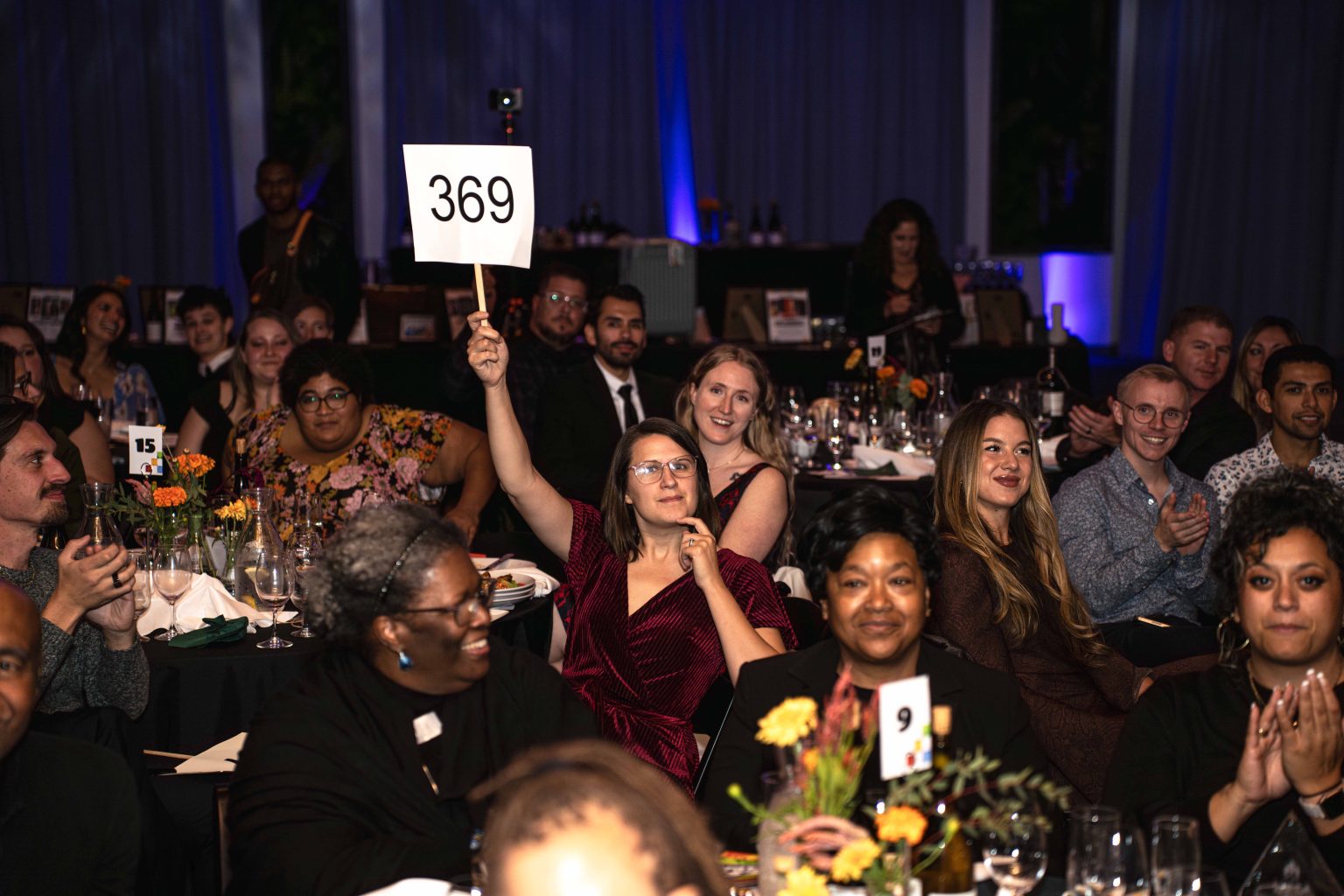 At the MESA Through the Decades 2025 Photo Gallery, a woman in a maroon dress sits at a round table, raising a sign with the number 369 as guests around her clap and smile in a warmly lit, festive room.