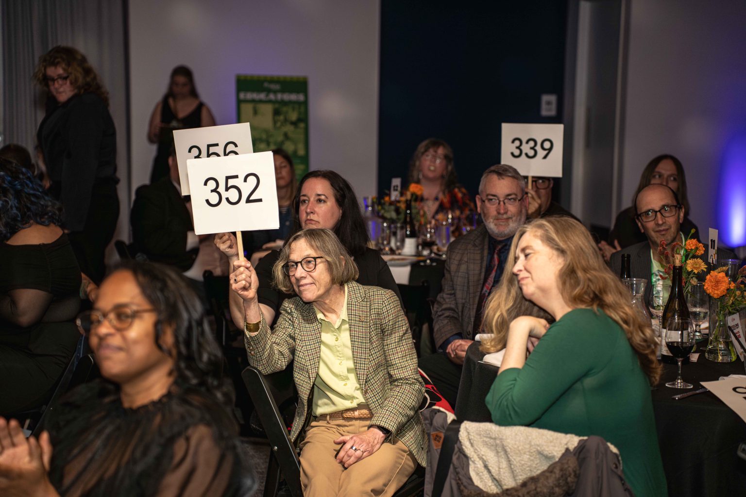 At the MESA Through the Decades 2025 Photo Gallery, people seated at a formal event hold up numbered paddles—352, 256, and 339—while attendees look toward the front, suggesting an auction or bidding event.