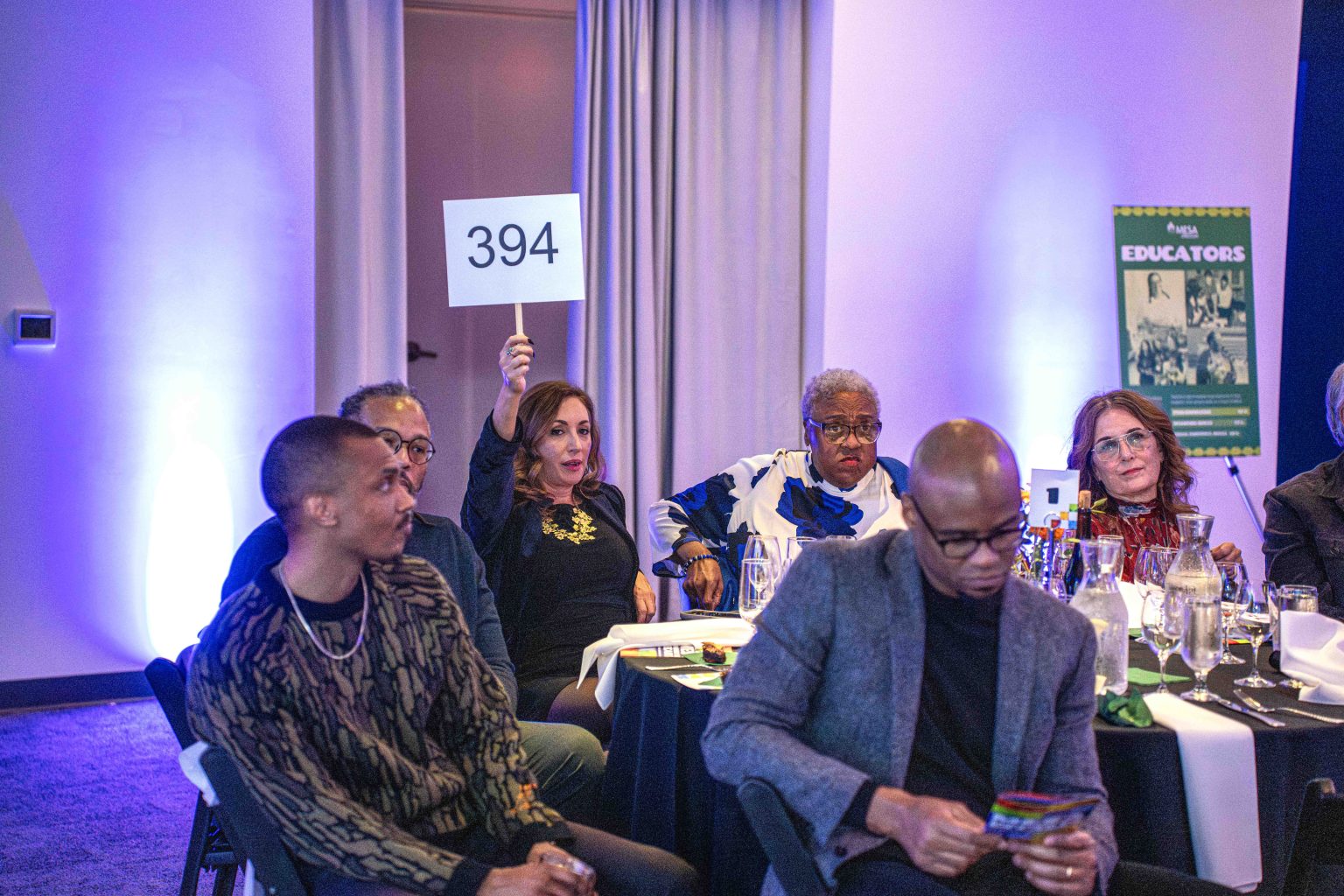 At the MESA Through the Decades 2025 Photo Gallery, a woman sits at a table holding up a sign with 394 as others watch attentively under purple lighting. A poster labeled EDUCATORS hangs on the wall in the background.