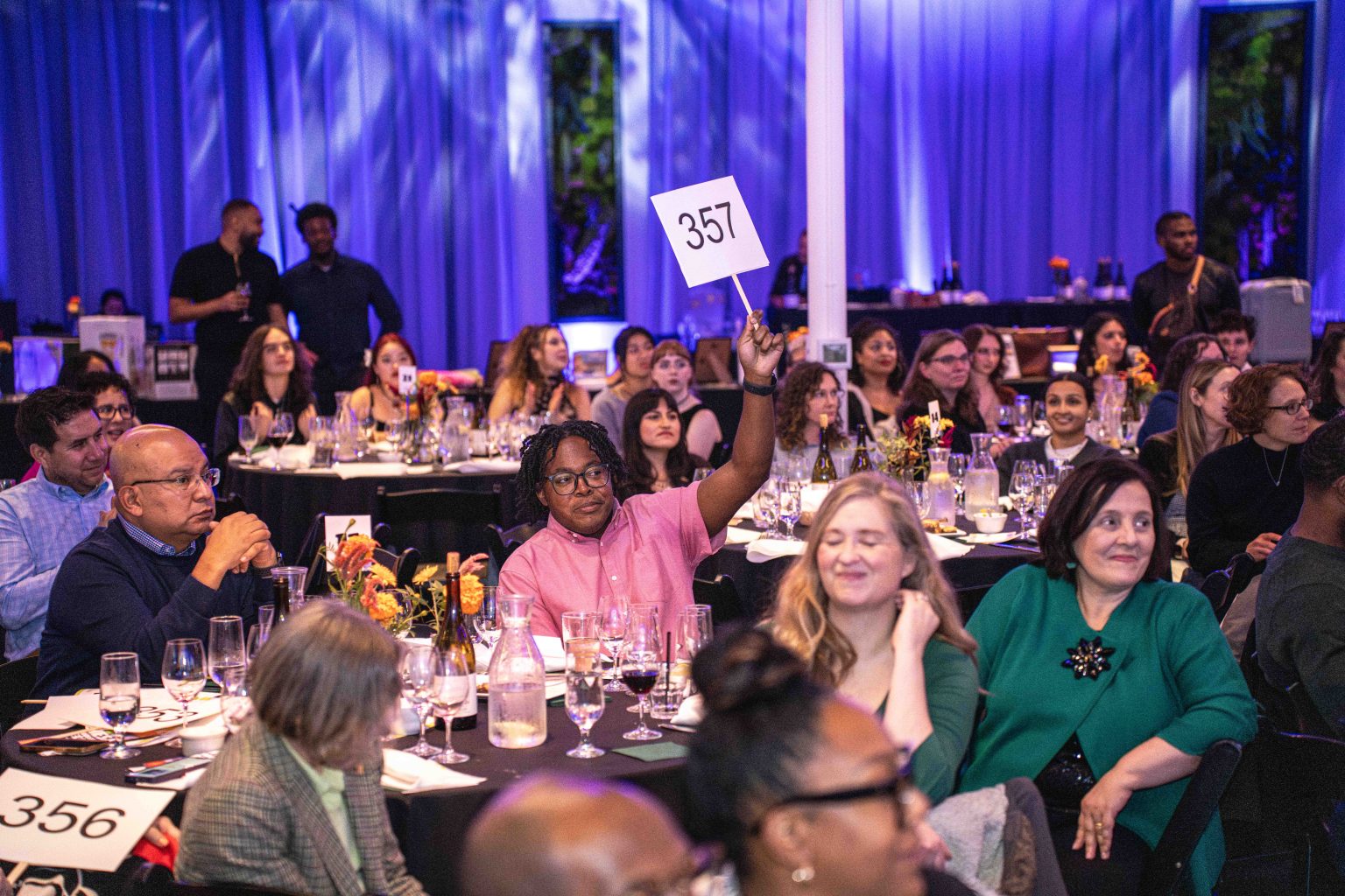 At a crowded auction event, part of the MESA Through the Decades 2025 Photo Gallery, a person in a pink shirt raises paddle 357 among attendees seated at flower-adorned tables under blue and purple lighting.