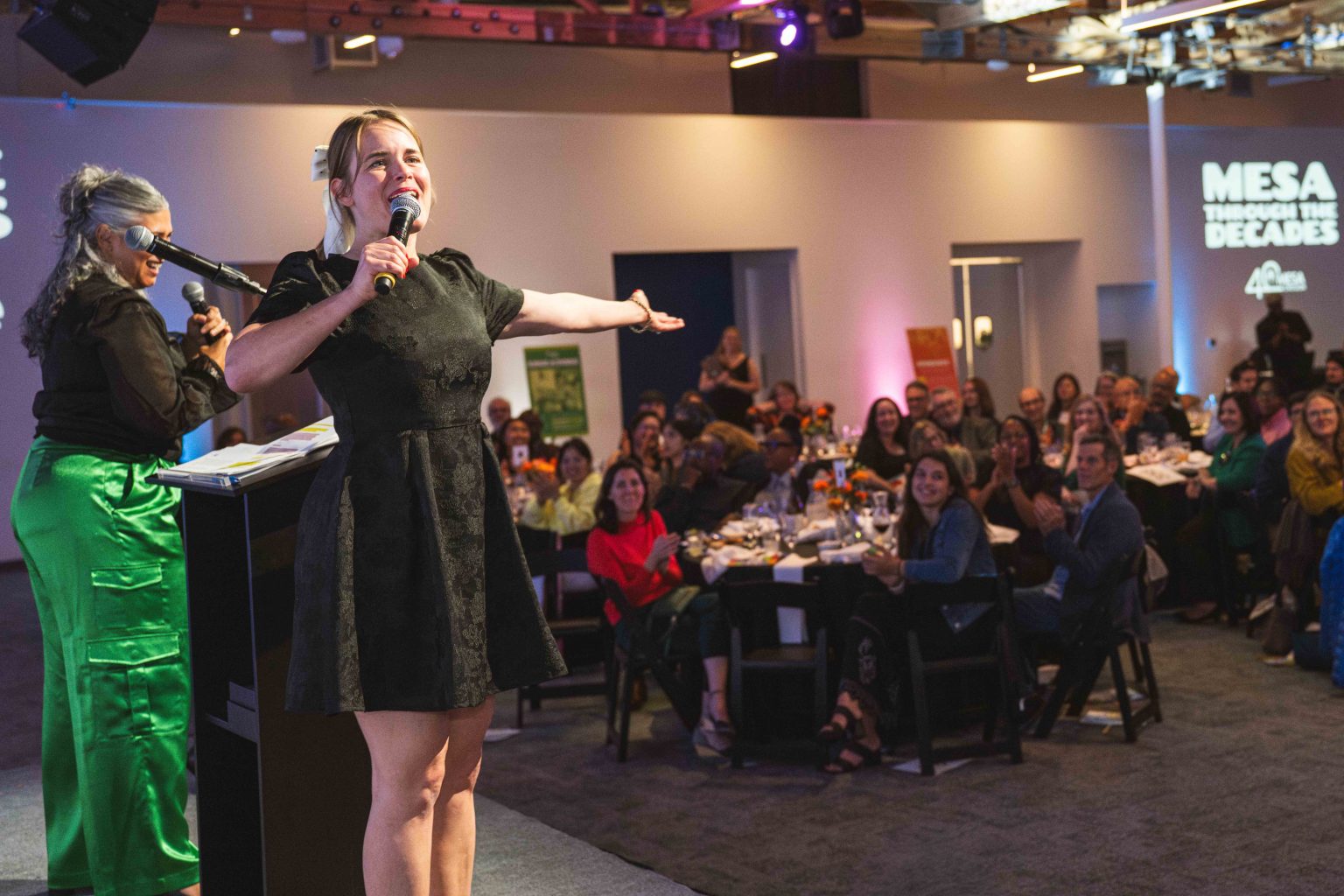 At the MESA Through the Decades 2025 Photo Gallery, a woman in a black dress speaks energetically on stage to an applauding audience, while another woman in a shiny green skirt stands at the podium with a microphone.