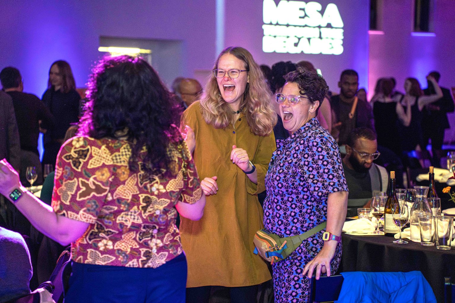 Three people stand and laugh together at a lively indoor event with colorful lighting. Other attendees mingle near tables with glasses and bottles. "MESA Through the Decades" is projected on a wall for the 2025 Photo Gallery.