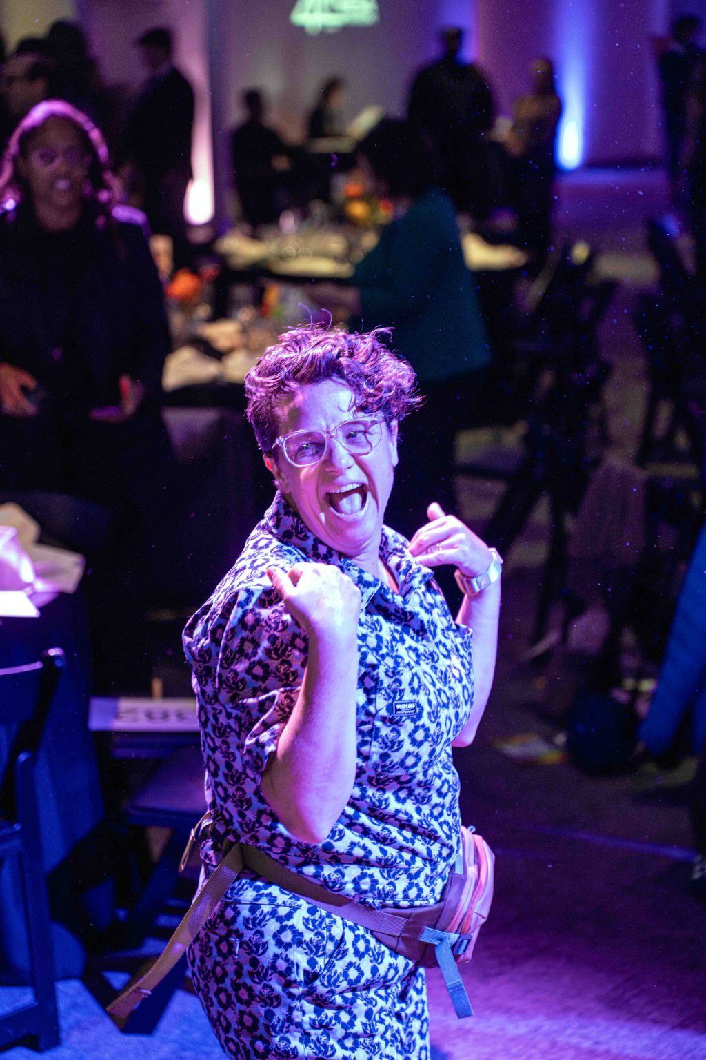 A person wearing glasses and a floral outfit smiles enthusiastically and points at themselves while posing in the lively, dimly lit MESA Through the Decades 2025 Photo Gallery, surrounded by people and vibrant purple lighting.