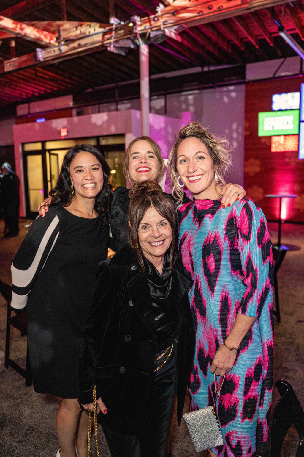 Four women smile and pose together at the MESA Through the Decades 2025 Photo Gallery event. Three stand at the back, one in front, with colorful lighting, exposed beams, and a few scattered chairs setting the vibrant scene.