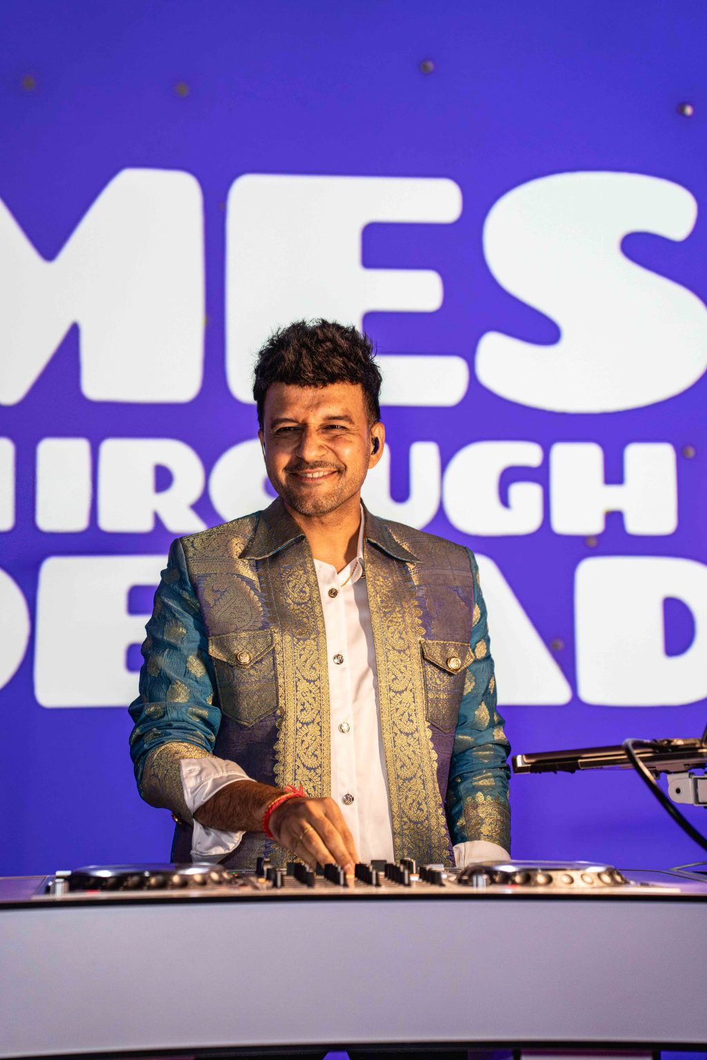 A man in a patterned blue and gold jacket is smiling while standing at a DJ console, performing in front of a bright purple background with large white text—part of the MESA Through the Decades 2025 Photo Gallery event.
