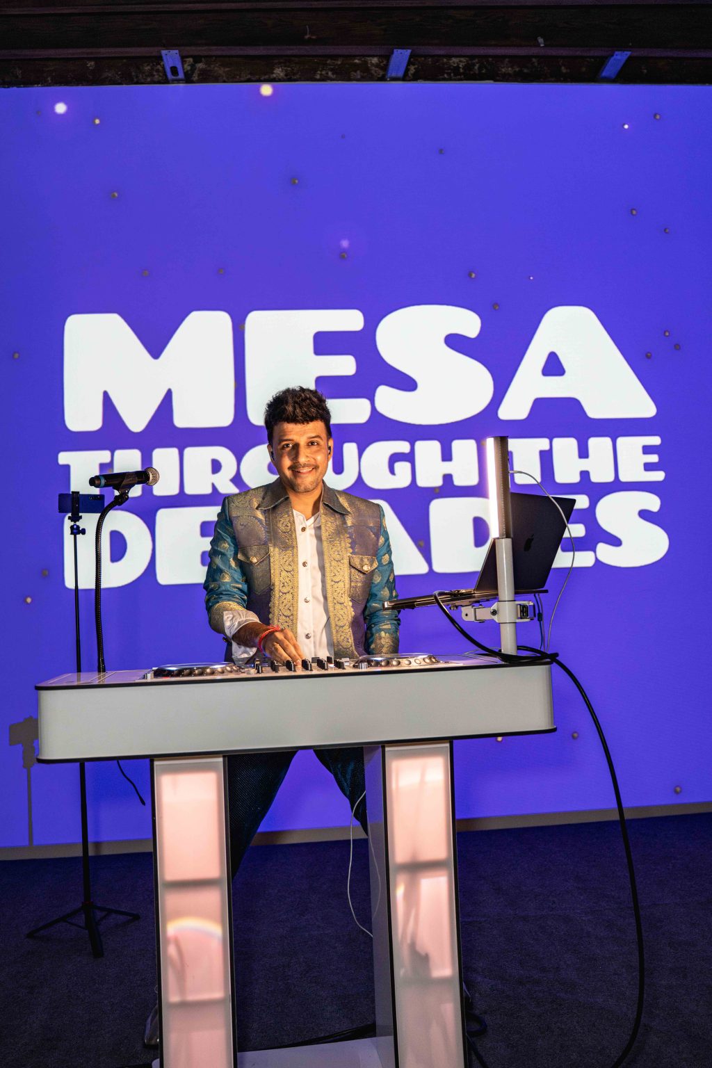 A DJ in a shiny blue and silver suit stands behind a modern DJ booth, smiling, with a purple-lit backdrop displaying the words MESA THROUGH THE DECADES—part of the MESA Through the Decades 2025 Photo Gallery.