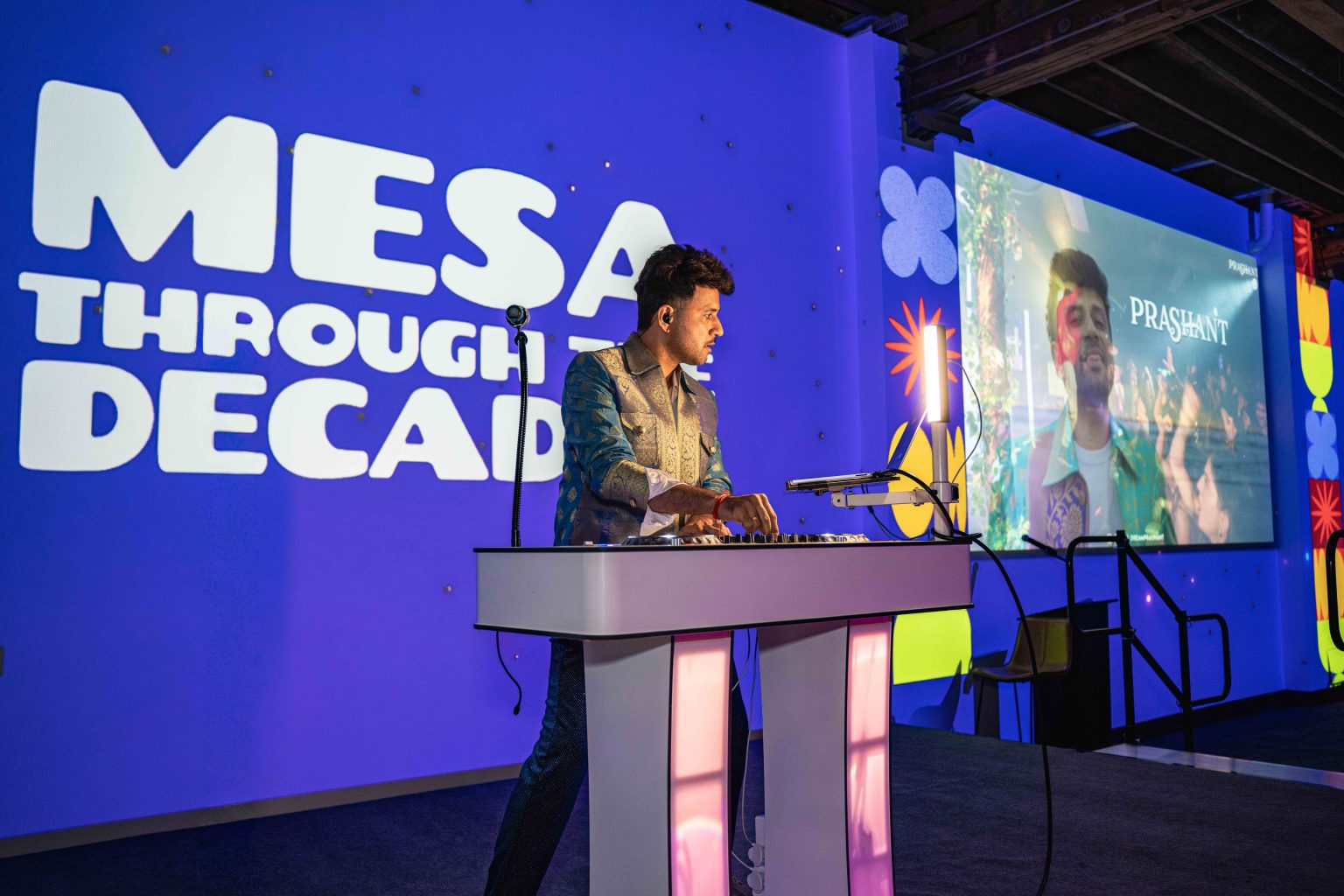 A man in a shiny suit plays keyboard on stage before a large screen reading “MESA THROUGH DECADES,” as vibrant graphics and another display showing a young man set the scene for the MESA Through the Decades 2025 Photo Gallery event.