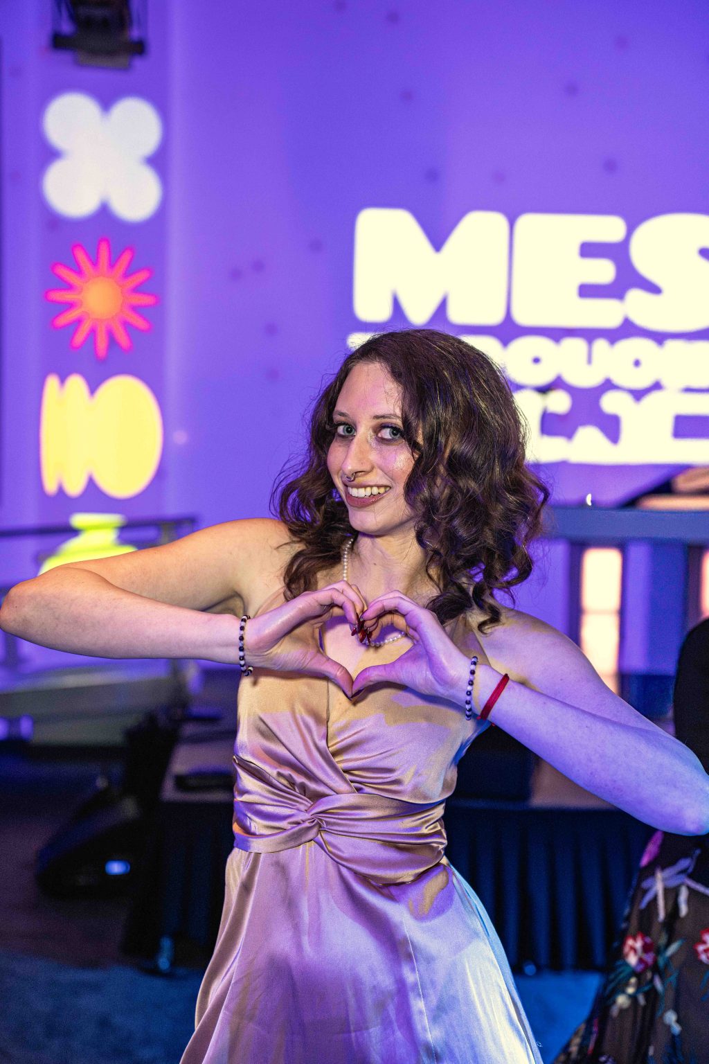 A woman in a shiny lavender dress stands indoors, smiling and forming a heart shape with her hands. Colorful, playful wall decorations and bright lights evoke a vibrant party mood at the MESA Through the Decades 2025 Photo Gallery.