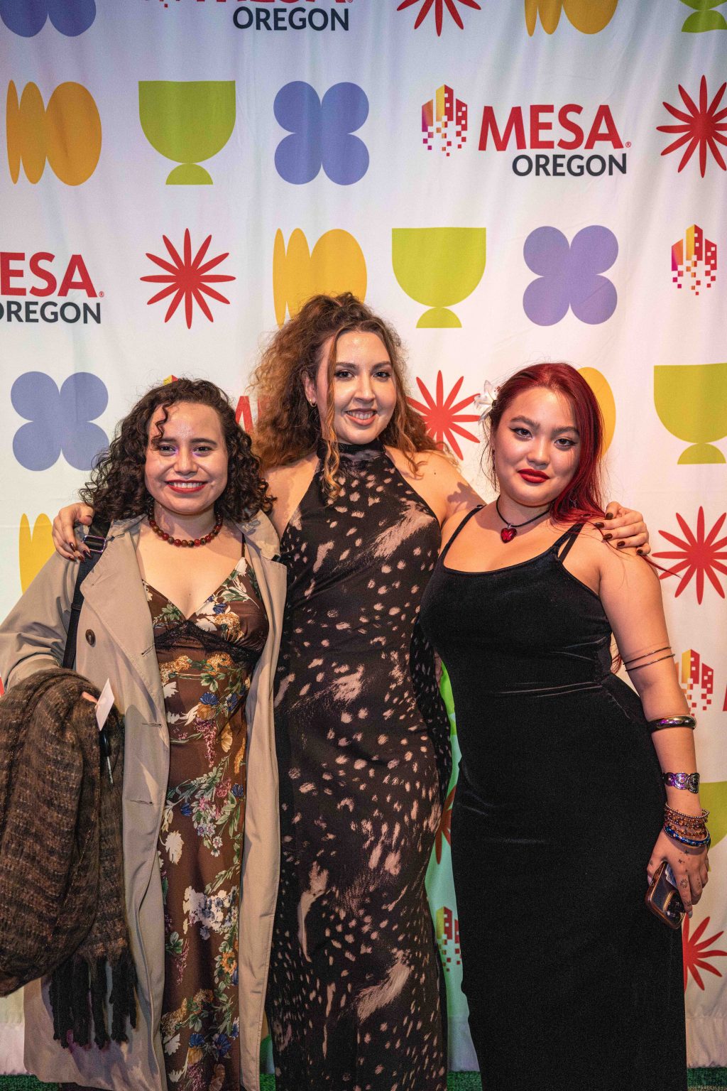 Three women in evening wear smile together in front of a colorful step-and-repeat banner featuring the MESA Oregon logo and geometric patterns at the MESA Through the Decades 2025 Photo Gallery.