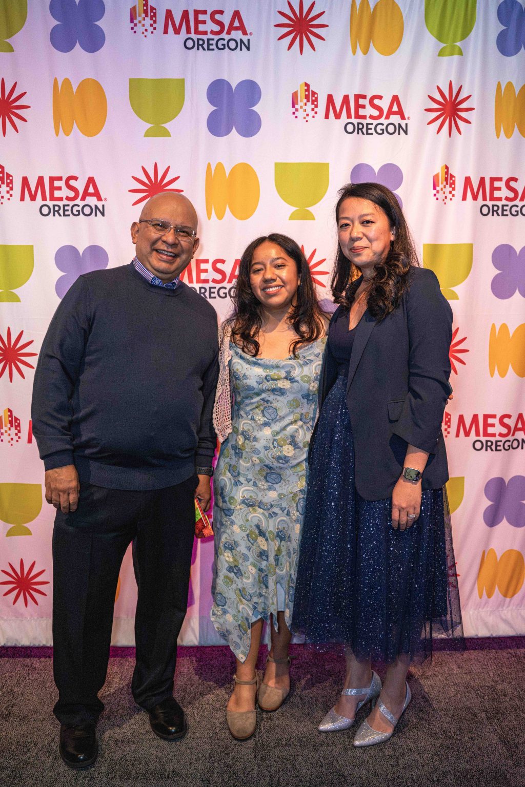 Three people stand smiling in front of a colorful MESA OREGON step-and-repeat banner at the MESA Through the Decades 2025 Photo Gallery, featuring geometric shapes in purple, yellow, orange, and green. Two women are in dresses, and one man wears a sweater and slacks.