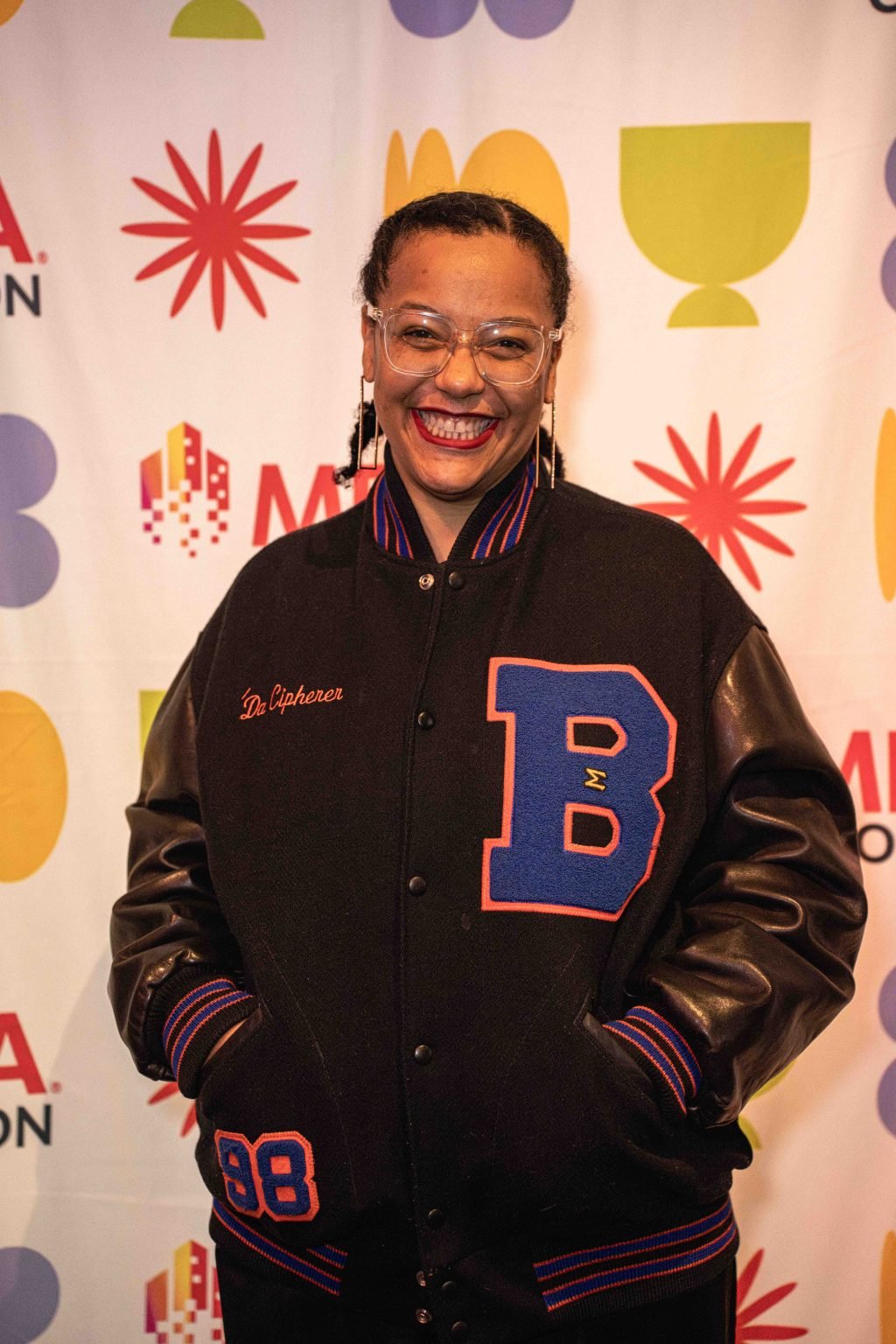 A smiling person with glasses and braided hair wears a black varsity jacket with a large blue B and Dr. Cipherer embroidered on it, standing in front of a colorful patterned backdrop at the MESA Through the Decades 2025 Photo Gallery.