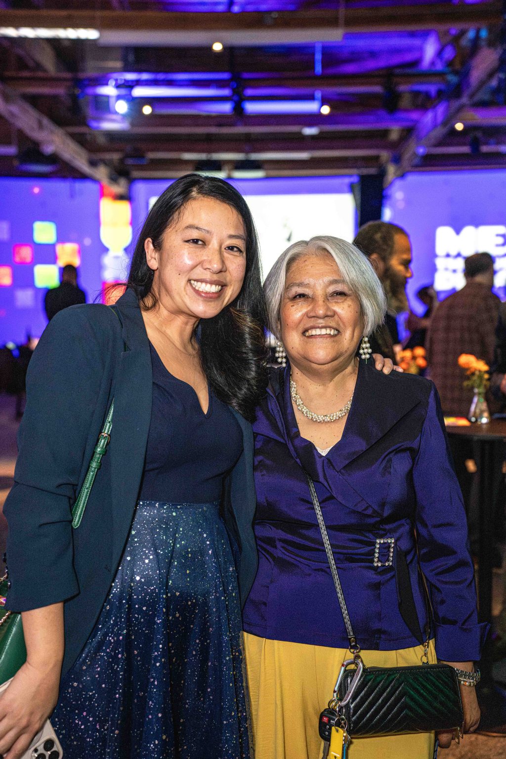 Two women smile and pose together at the MESA Through the Decades 2025 Photo Gallery event, dressed formally amid purple lighting, colorful lights, a large screen, and fellow attendees in the background.