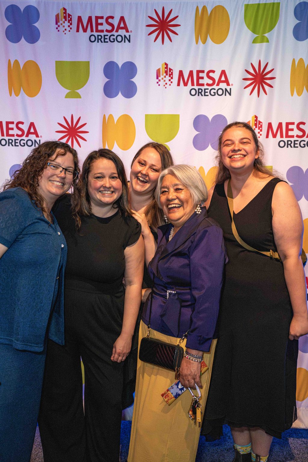 Five women stand smiling together in front of a colorful MESA Oregon backdrop, dressed in semi-formal attire, posing closely for a group photo at the MESA Through the Decades 2025 Photo Gallery event.