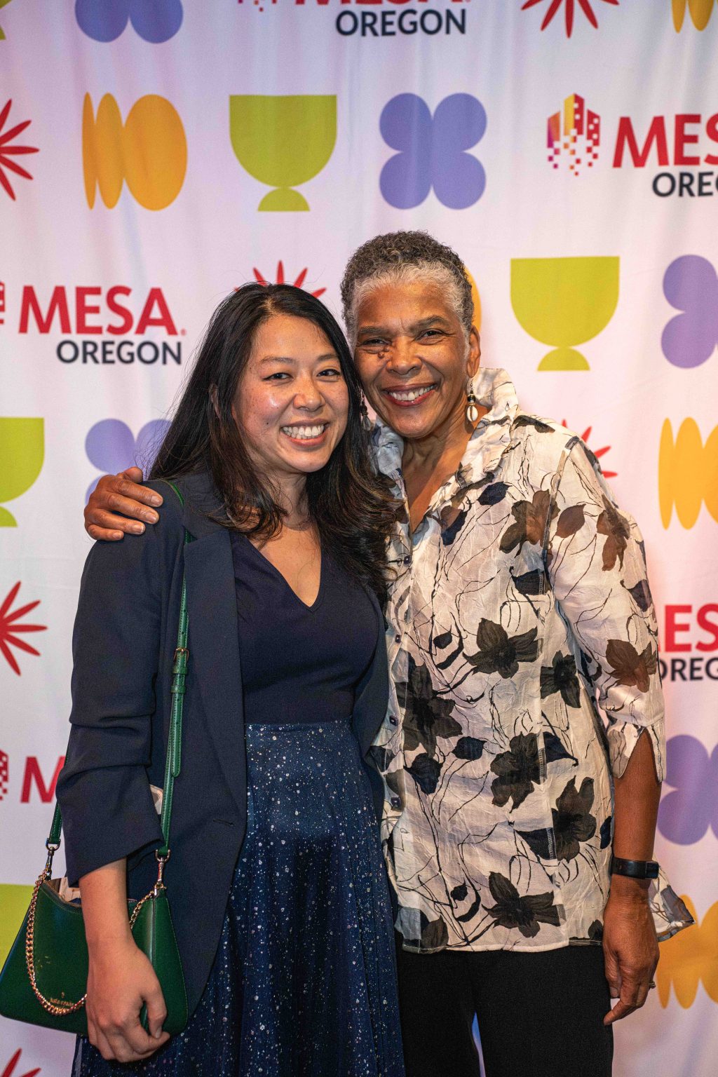 Two women smiling and posing together in front of a colorful step-and-repeat backdrop with Mesa Oregon logos and abstract shapes at the MESA Through the Decades 2025 Photo Gallery. One woman has her arm around the other.