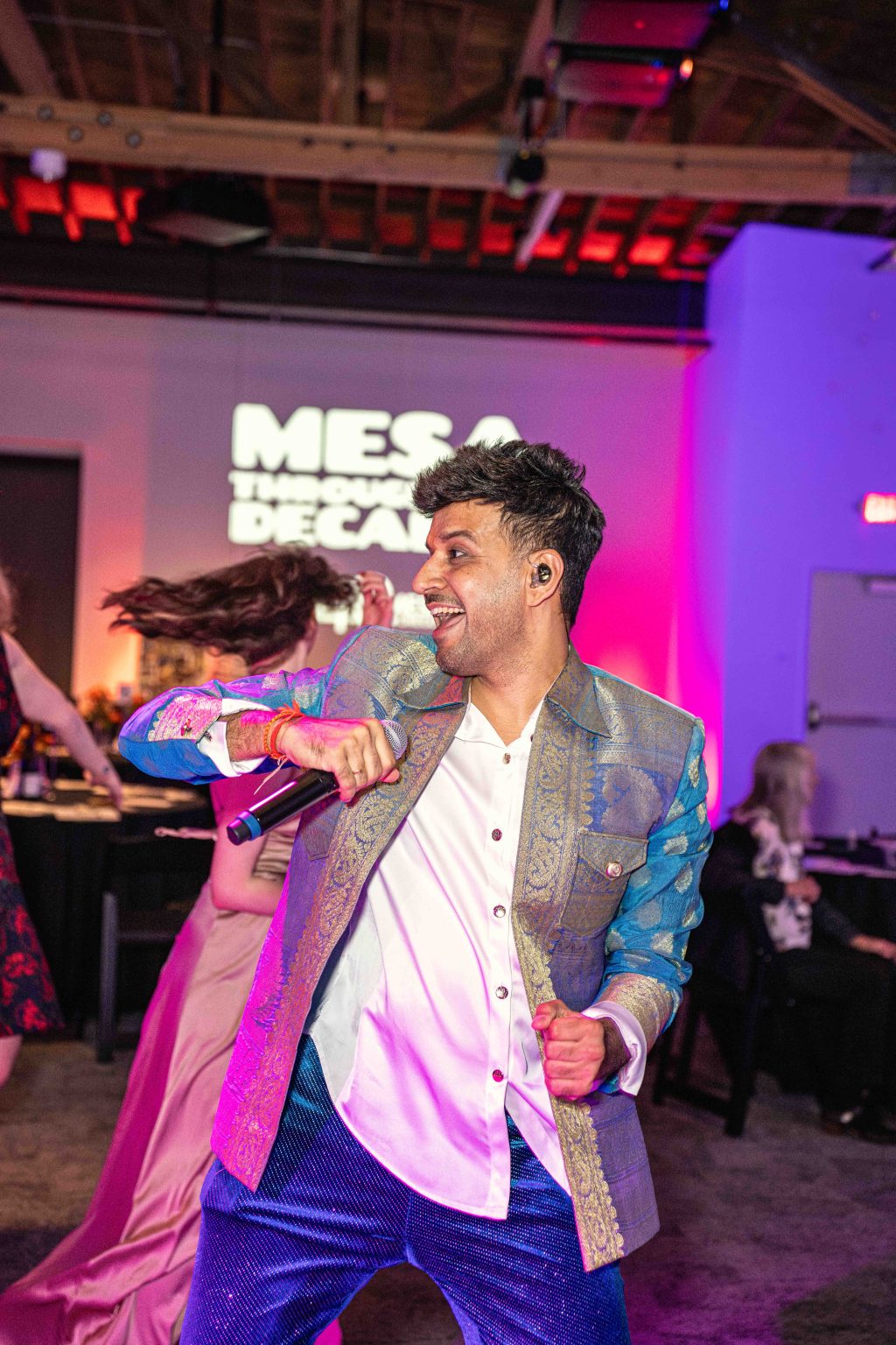 A man in a colorful suit sings energetically into a microphone on stage, as people dance behind him under vibrant purple and pink lighting at the MESA Through the Decades 2025 Photo Gallery indoor event.
