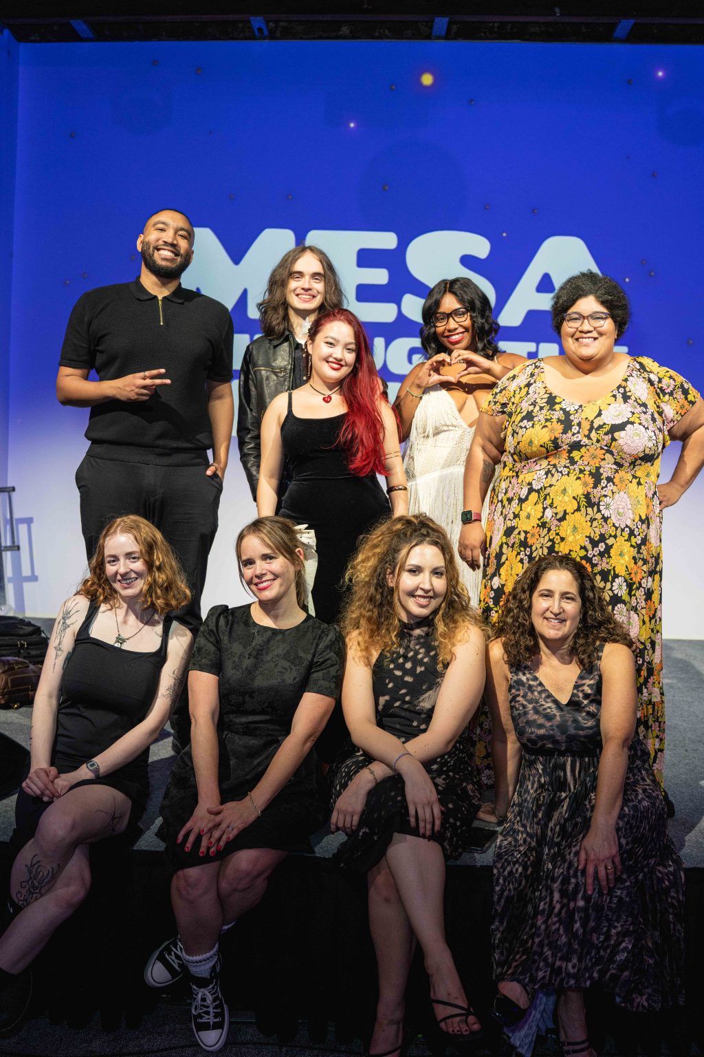 A group of nine people pose and smile together on a stage in front of a blue and white MESA sign for the MESA Through the Decades 2025 Photo Gallery. They are dressed in a variety of outfits, with some sitting and some standing.