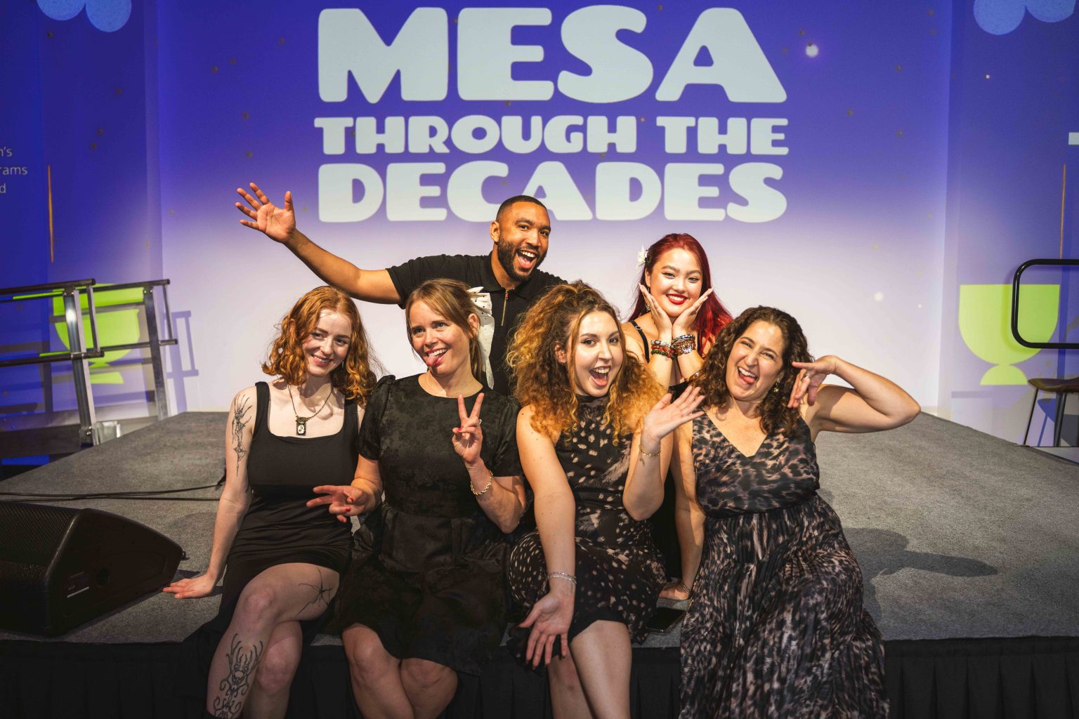 Six people pose enthusiastically on stage, smiling and making playful gestures in front of a lit screen that reads MESA THROUGH THE DECADES—captured perfectly for the MESA Through the Decades 2025 Photo Gallery.