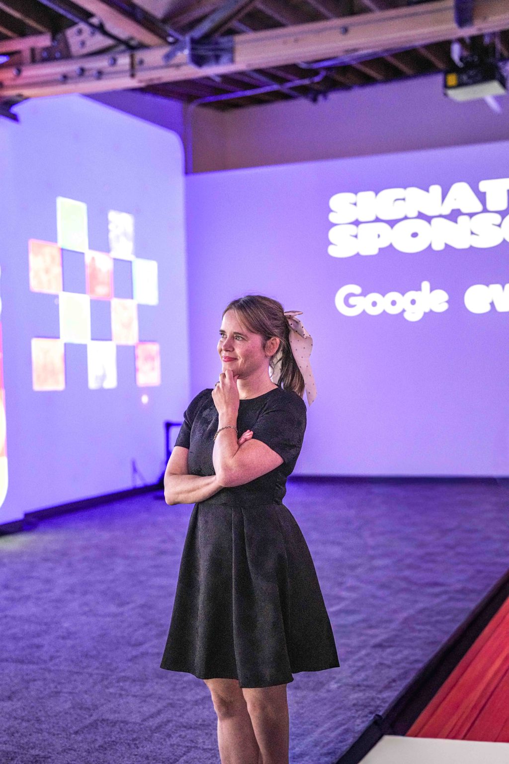 A woman in a black dress stands thoughtfully in a modern, purple-lit room with projected displays on the walls, including the text SIGNAT SPONS and Google ev, part of the MESA Through the Decades 2025 Photo Gallery.