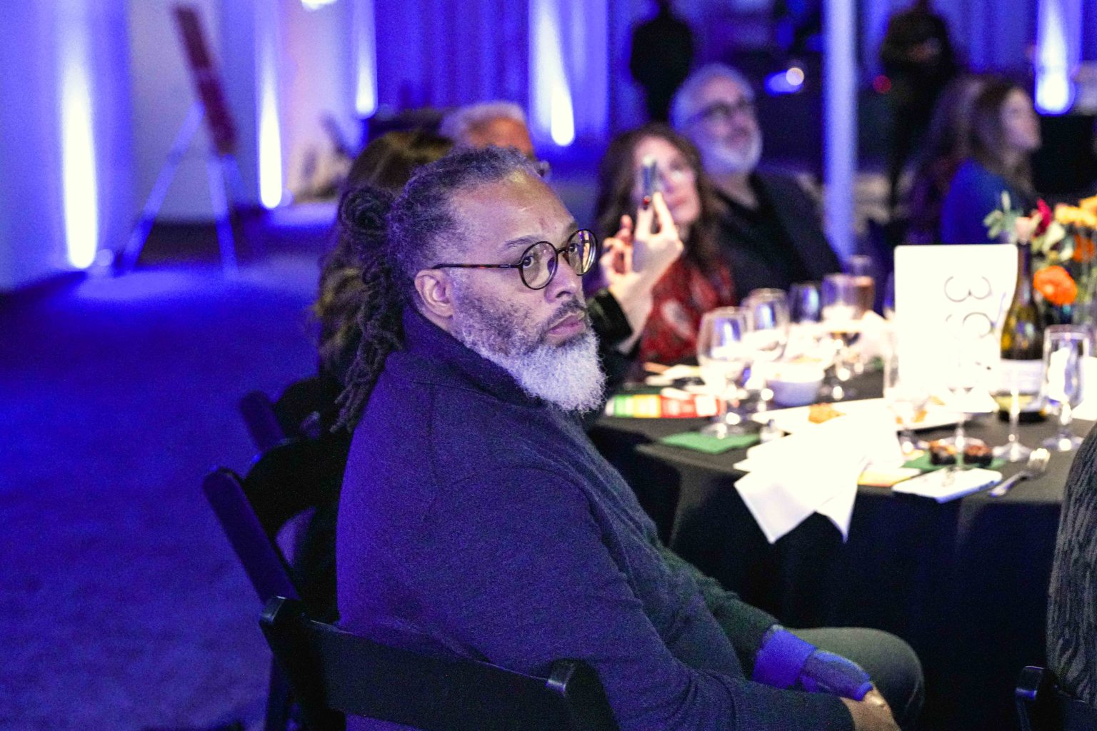 At the MESA Through the Decades 2025 Photo Gallery, a man with gray hair and a beard sits at a round table, surrounded by others. The softly lit room glows with blue lights, and tables are adorned with glasses, plates, and flowers.