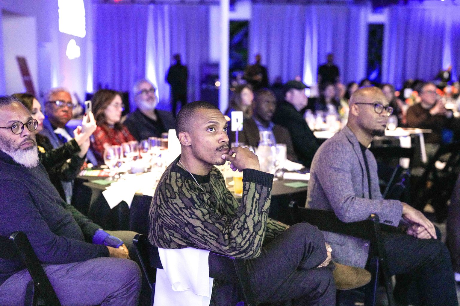 Guests at the MESA Through the Decades 2025 Photo Gallery sit at round tables in a dimly lit event space, attentively watching something off-camera. The blue and purple ambiance and decorated tables set a stylish tone for this semi-formal gathering.