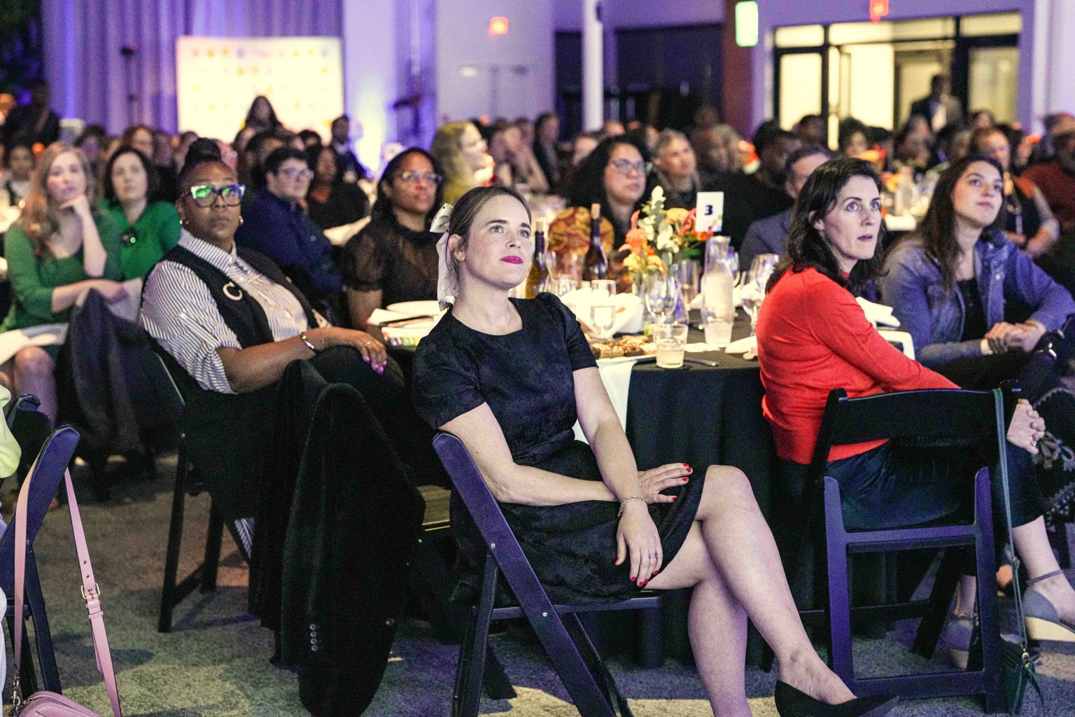 A diverse group of people sit at round tables, attentively watching something off-camera at a formal indoor event. The scene, part of the MESA Through the Decades 2025 Photo Gallery, features flowers, drinks, and papers on each table.
