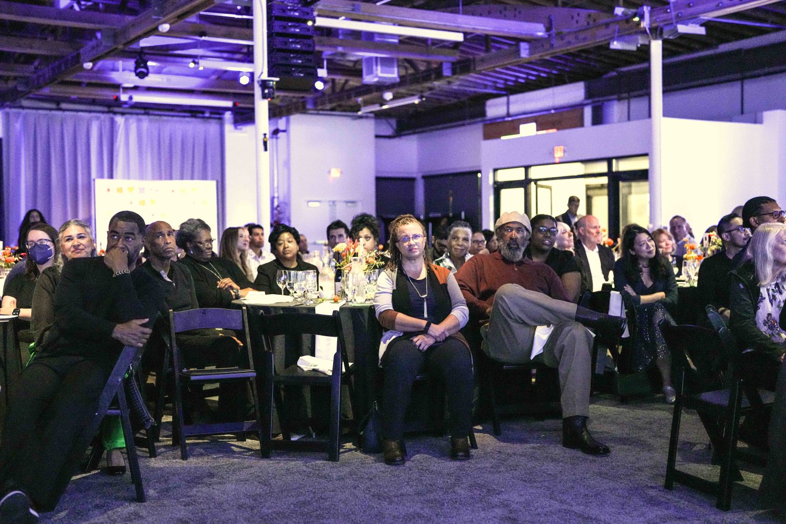 A diverse group of people sit at tables in a large, modern event space during the MESA Through the Decades 2025 Photo Gallery, attentively watching something off-camera. The room is warmly lit with purple accents and decorated with flowers.