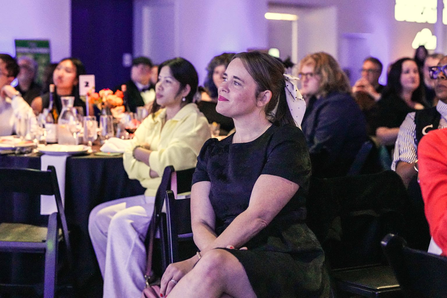 A woman in a black dress with a white hair ribbon sits and smiles while watching an event at the MESA Through the Decades 2025 Photo Gallery, surrounded by other attendees seated at round tables in a softly lit room.