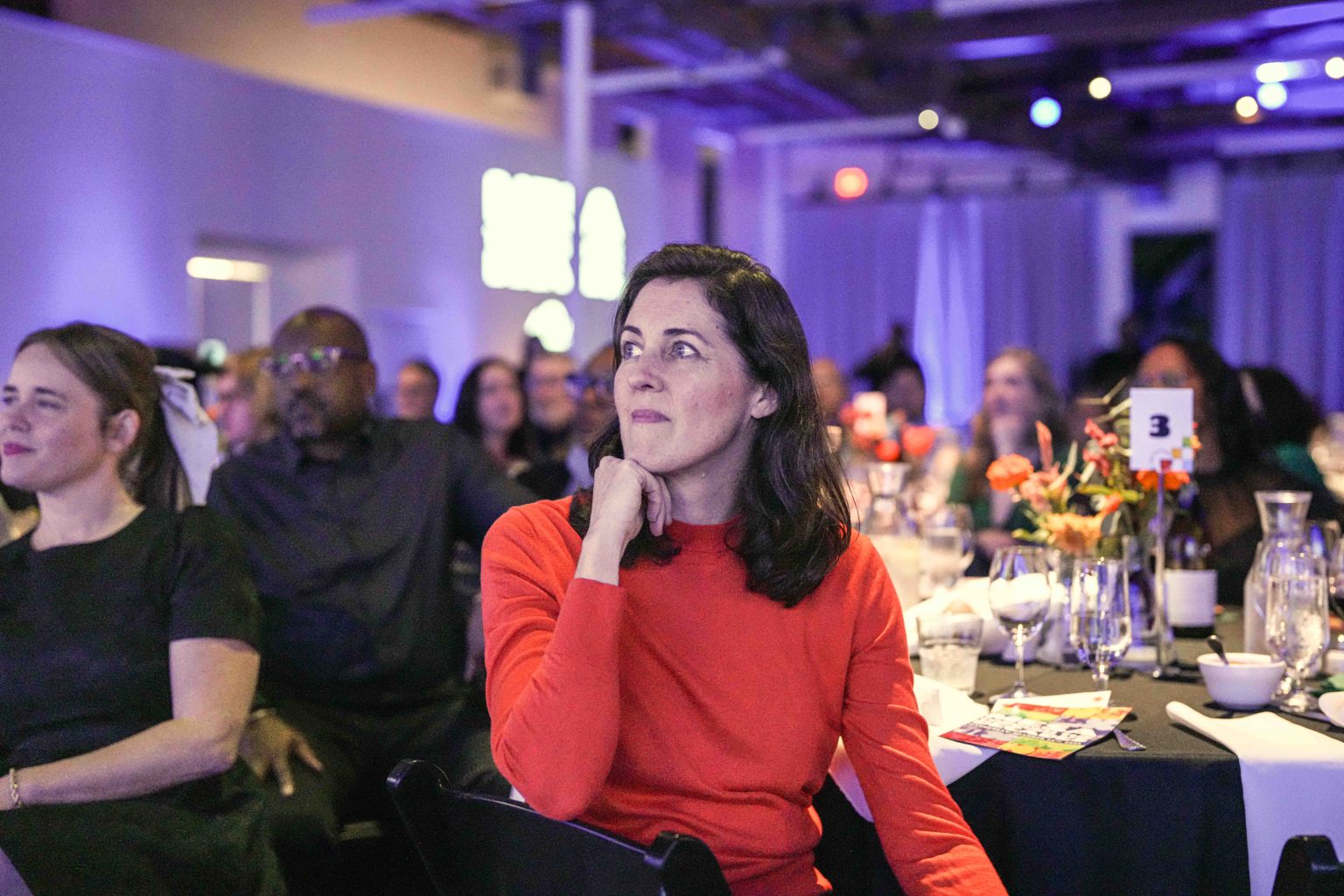 A woman in a red top sits at a table, resting her chin on her hand, attentively watching an event at the MESA Through the Decades 2025 Photo Gallery in a dimly lit venue filled with people and round tables decorated with flowers.