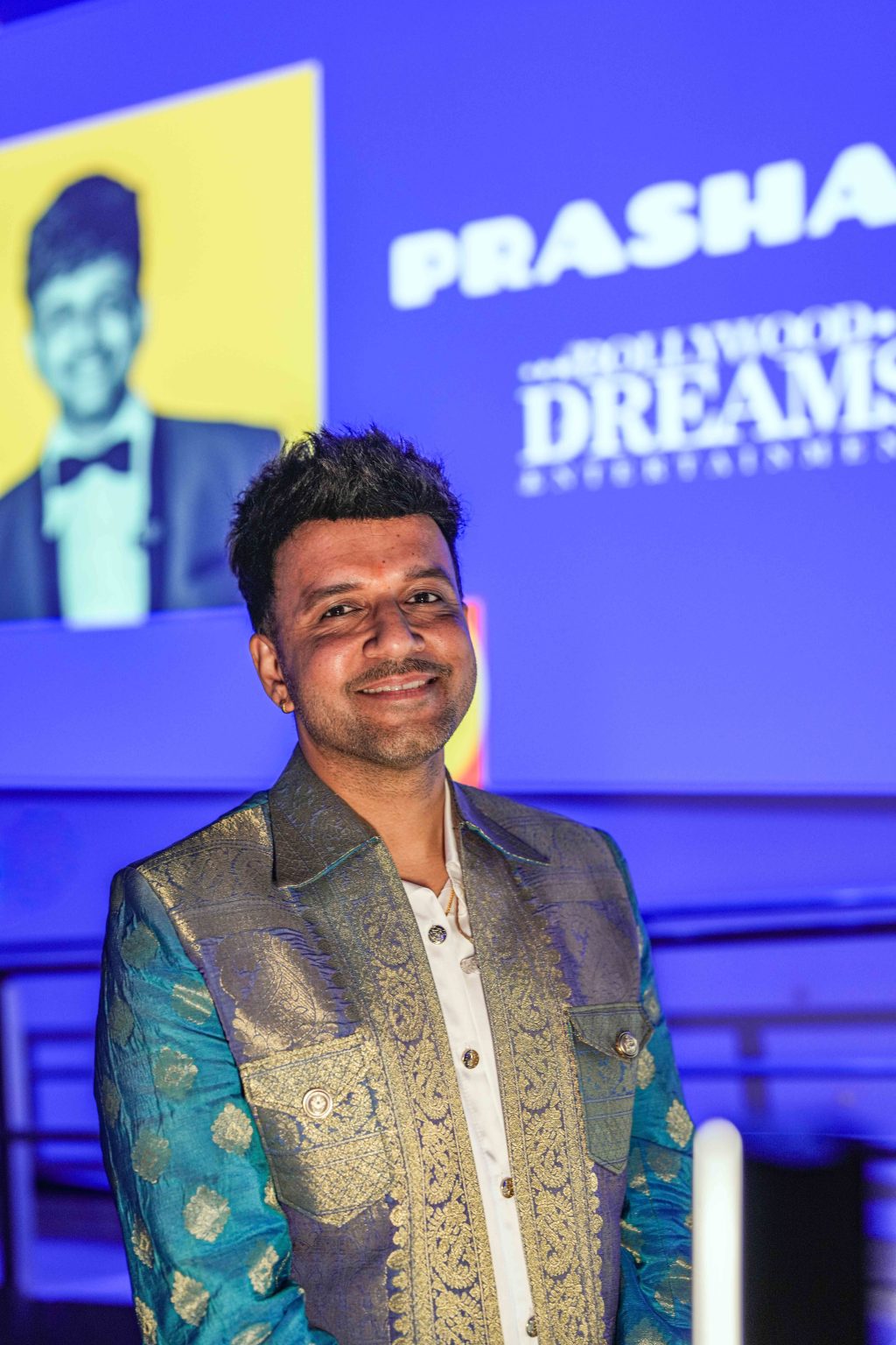 A man in a blue and gold embroidered jacket smiles at the camera, standing in front of a blue-lit background with a screen displaying a black-and-white portrait and the words Prasha and Dreams at the MESA Through the Decades 2025 Photo Gallery.