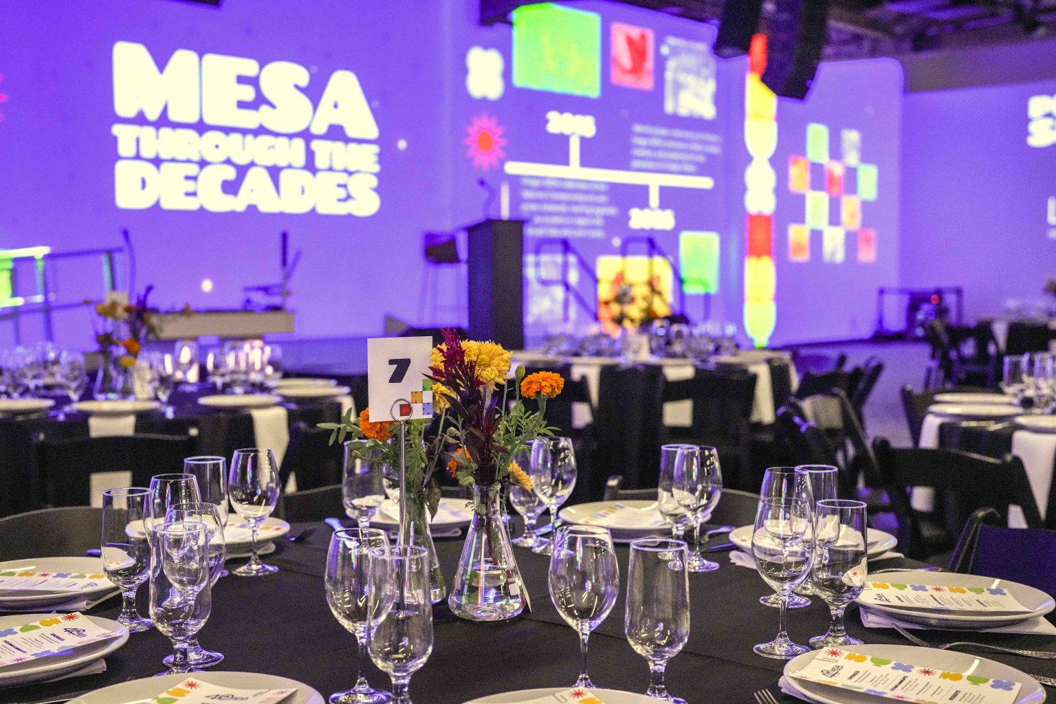 A banquet hall with round tables set for dinner, featuring glassware, menus, and floral centerpieces. In the background, a large wall display reads “MESA Through the Decades” and spotlights scenes from the 2025 Photo Gallery timeline.