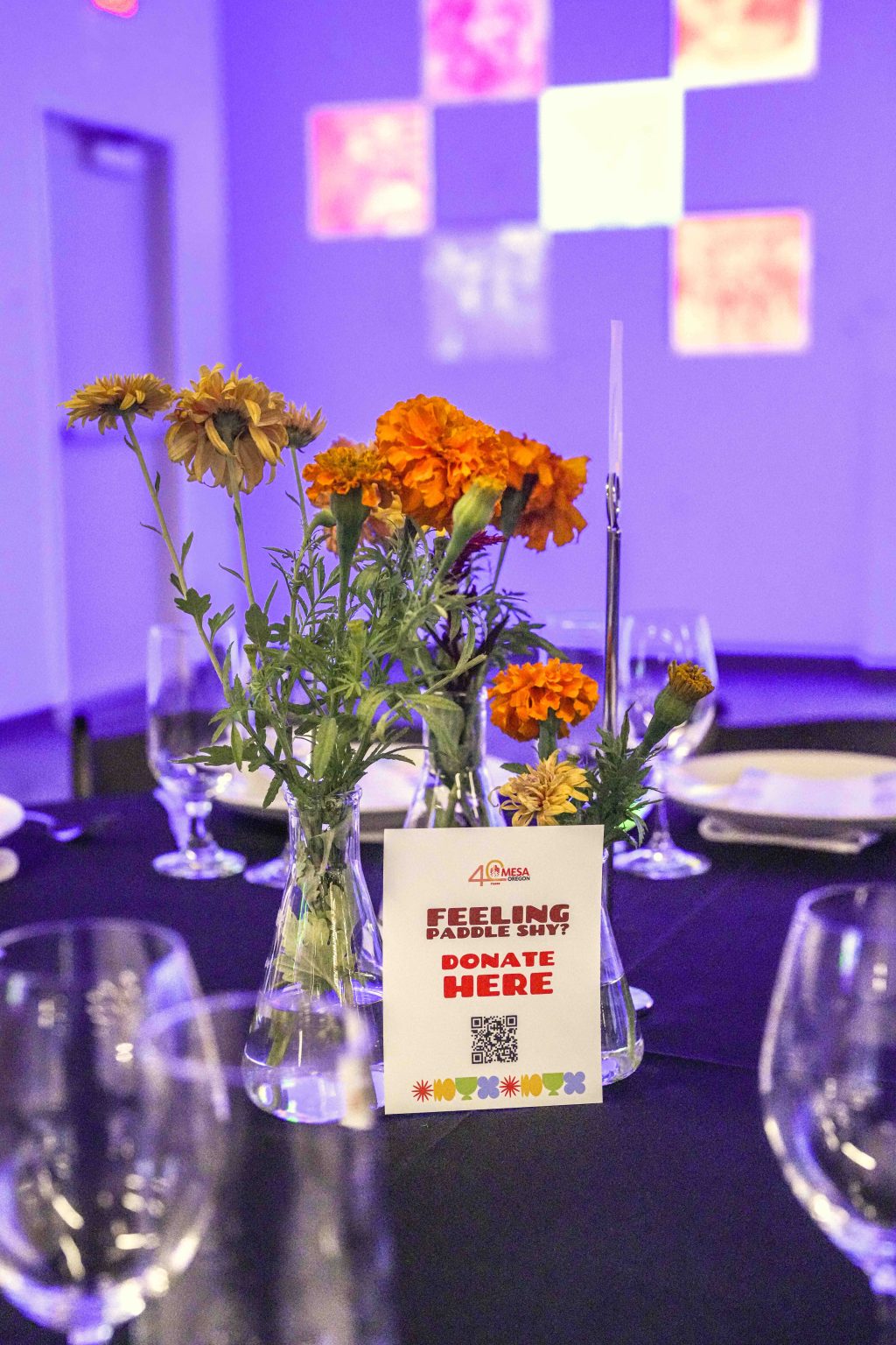 A table centerpiece with yellow and orange flowers in glass vases and a donation sign with a QR code sits on a black tablecloth, surrounded by empty wine glasses and plates. The softly lit purple background highlights the MESA Through the Decades 2025 Photo Gallery theme.
