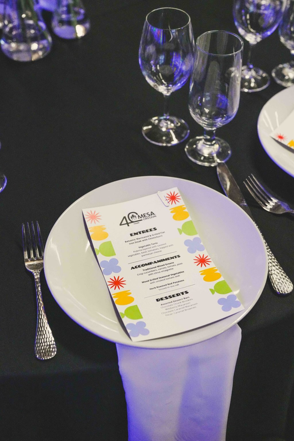 A white dinner plate with a colorful printed menu rests on a black tablecloth, flanked by a fork and knife. In the background, empty glasses and another place setting evoke the elegance of the MESA Through the Decades 2025 Photo Gallery.