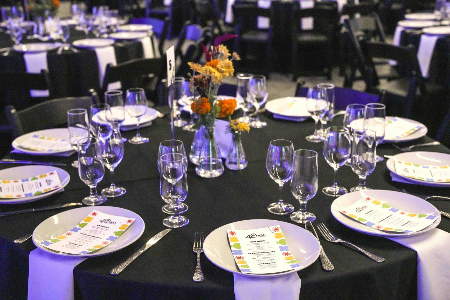 A round dinner table set for an event with plates, menus, glassware, cutlery, and a small centerpiece of colorful flowers—perfect for capturing in the MESA Through the Decades 2025 Photo Gallery—surrounded by black chairs and similar tables in the background.