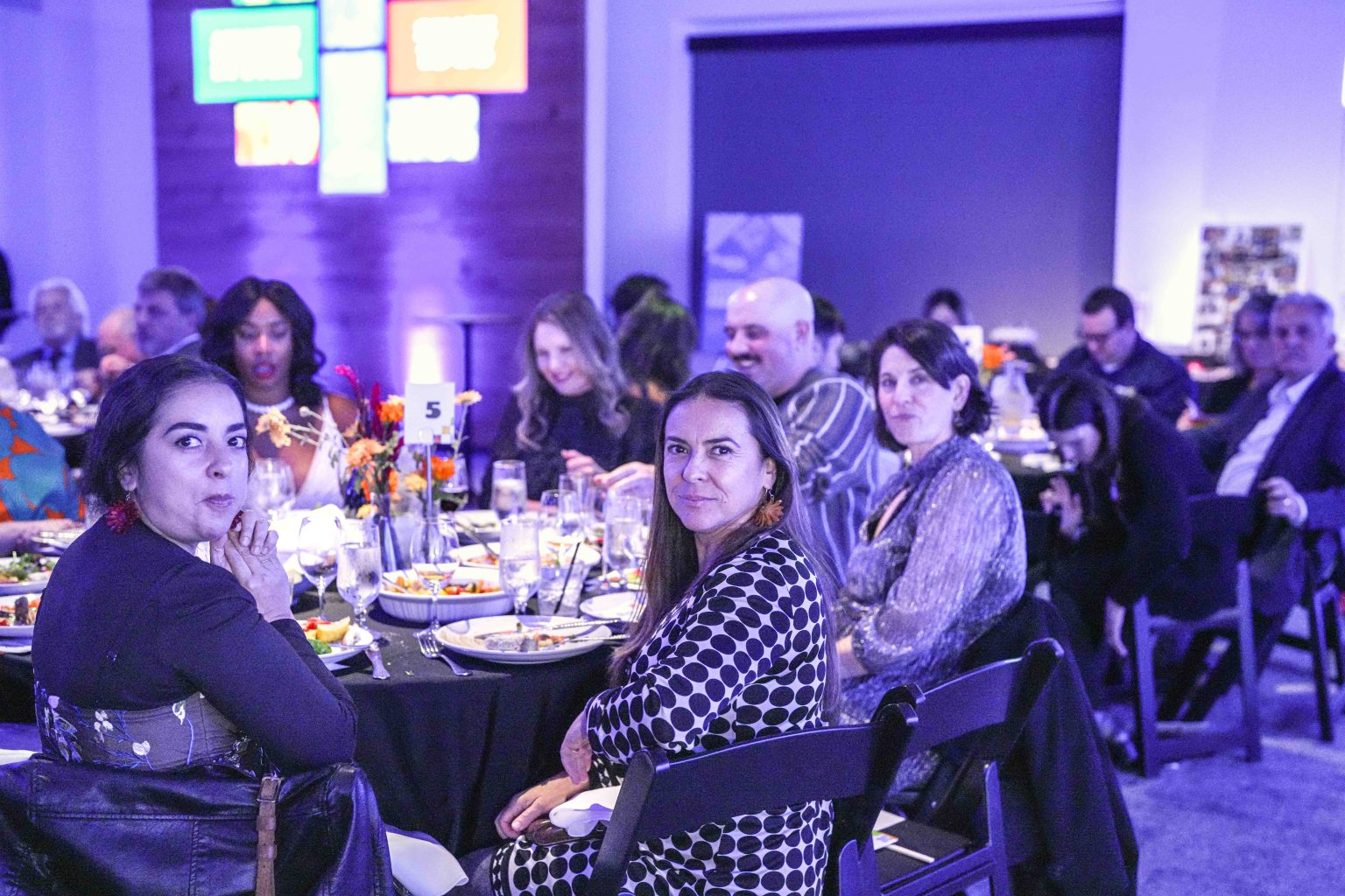People sit around tables at a formal event, looking toward the camera in the dimly lit, blue-hued room. Plates of food and drinks fill the tables while colorful signs decorate the wall, capturing a special moment for the MESA Through the Decades 2025 Photo Gallery.