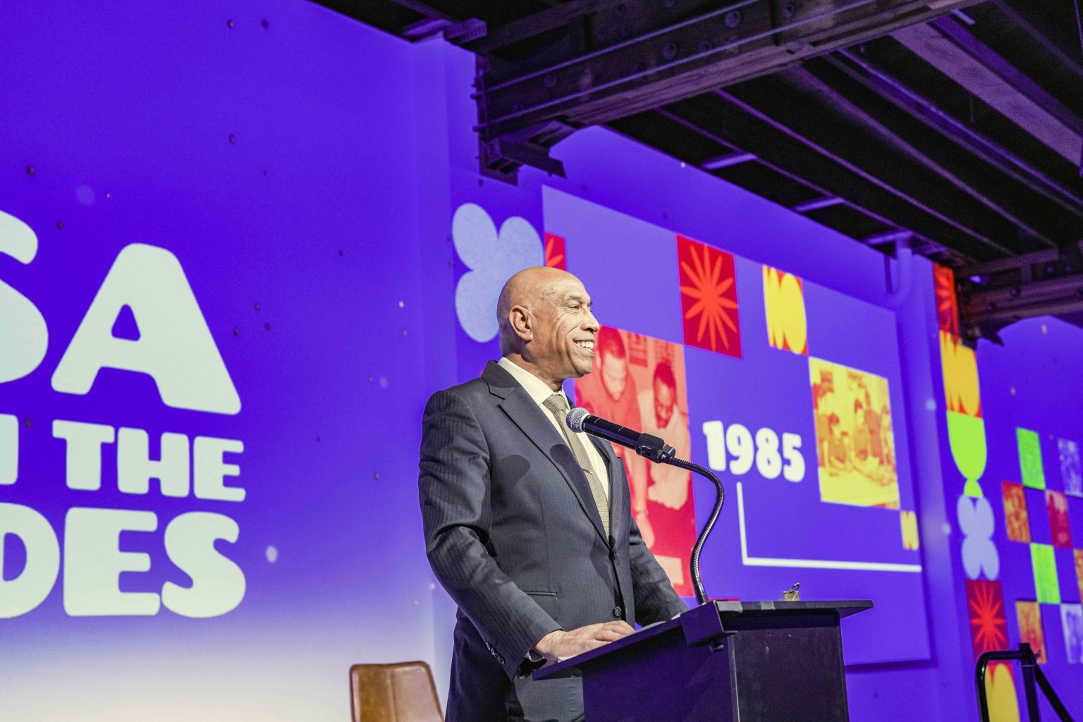 A man in a suit stands and smiles at a podium with a microphone, speaking at an event. The background displays colorful graphics and the text “1985,” “USA IN THE 80S,” and "MESA Through the Decades 2025 Photo Gallery" on a purple-lit wall.