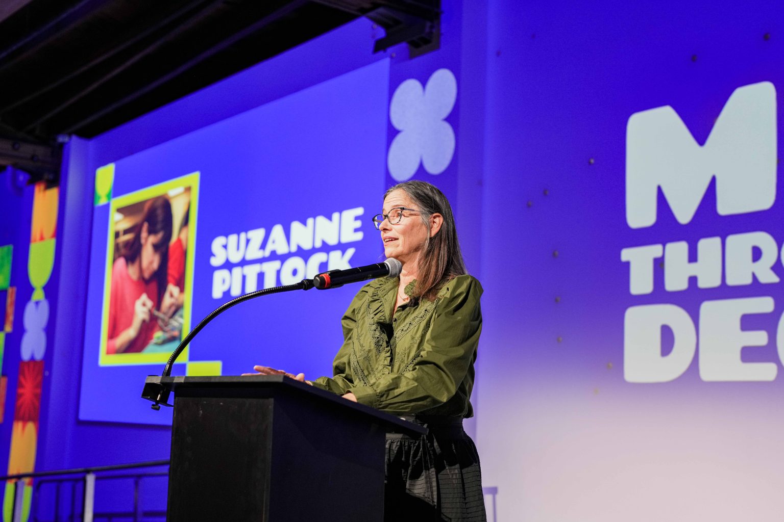 A woman stands at a podium speaking into a microphone. Behind her, a screen displays the name Suzanne Pittock, her photo, and colorful graphics on a purple background from the MESA Through the Decades 2025 Photo Gallery.