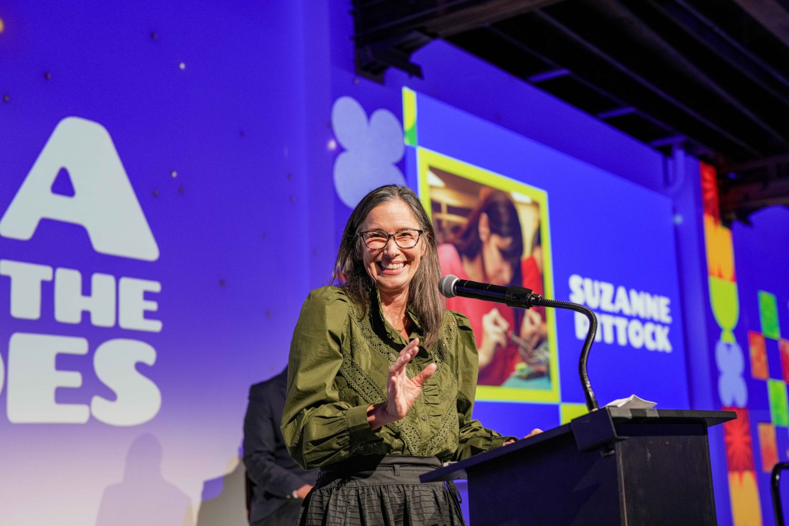 A woman with long dark hair and glasses smiles while speaking at a podium. Behind her, the MESA Through the Decades 2025 Photo Gallery displays a photo and the name Suzanne Dottock in a brightly lit event space.