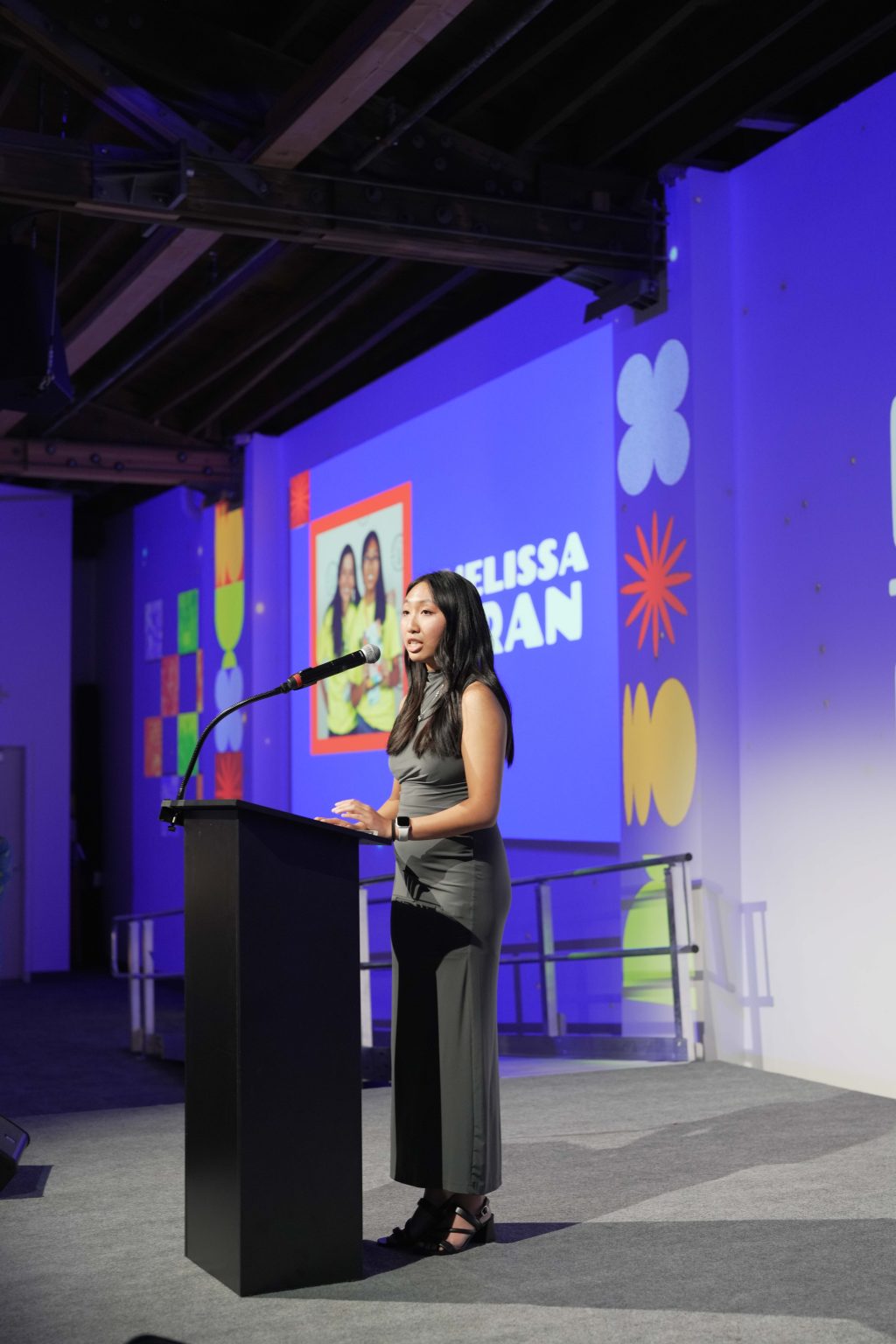 A woman stands at a podium speaking into a microphone on stage with a blue backdrop displaying colorful shapes, the name Melissa Tran, and "MESA Through the Decades 2025 Photo Gallery." A photo of three women is projected behind her.