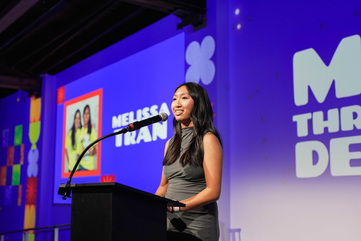 A woman stands at a podium speaking into a microphone. Behind her, a large screen displays her picture and the name Melissa Tran along with colorful designs and text for the MESA Through the Decades 2025 Photo Gallery event or presentation.