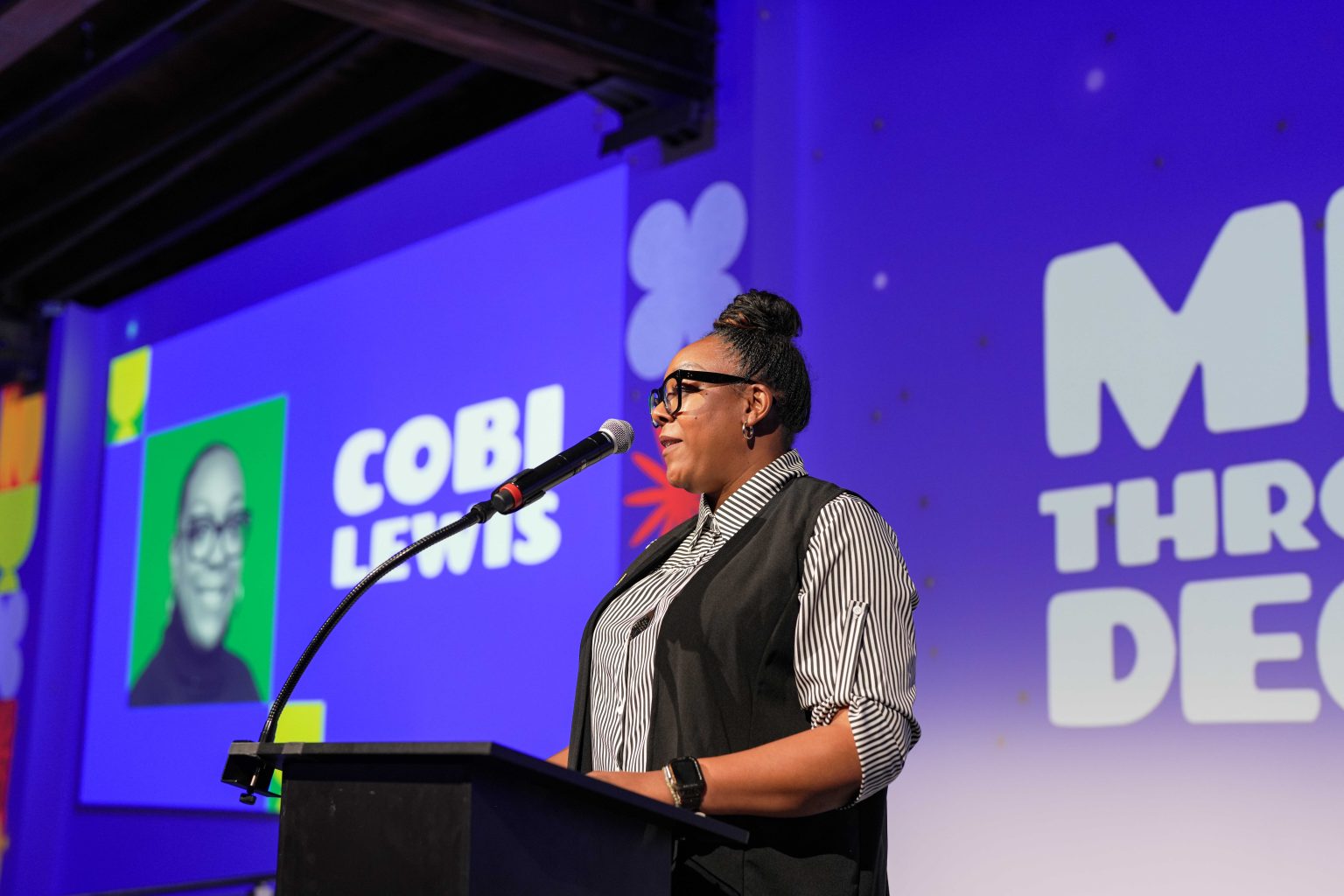 A person wearing glasses speaks at a podium with a microphone. Behind them, a large screen displays the name Cobi Lewis, a black-and-white portrait, colorful graphics, and the phrase "MOVE THROUGH DEC" from the MESA Through the Decades 2025 Photo Gallery.