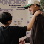 Two students stand in front of a science fair display titled “Ponchos” at the 2026 Oregon MESA Demo Days. One smiles and pats the other’s shoulder as they talk, with project posters and diagrams visible in the background.